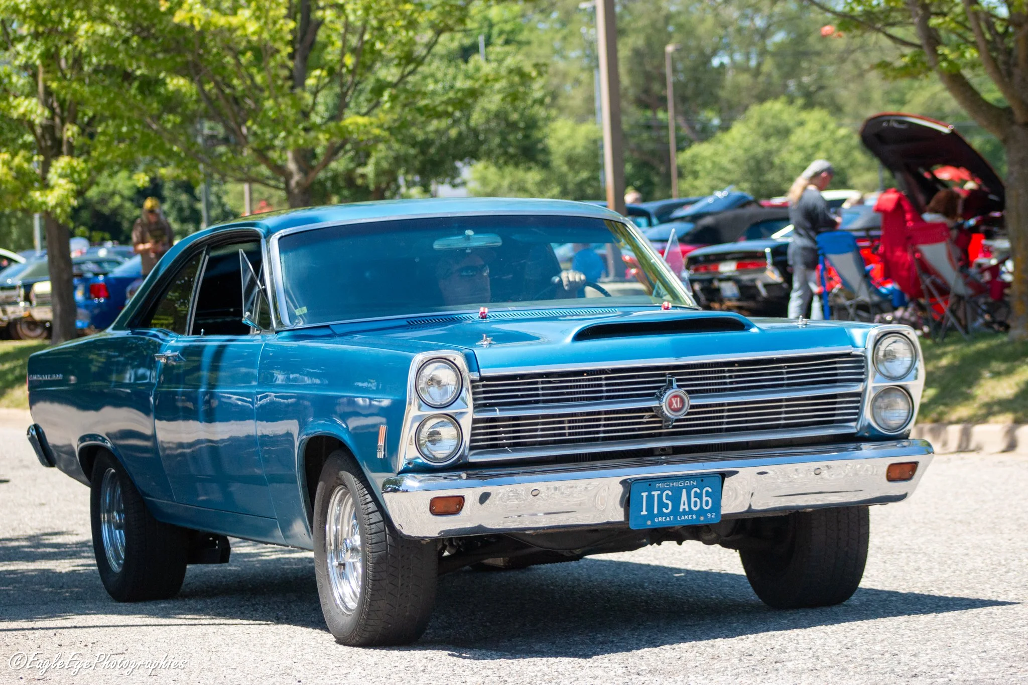 A vintage blue car at an outdoor car show, with other classic cars and people in the background, shaded by trees.
