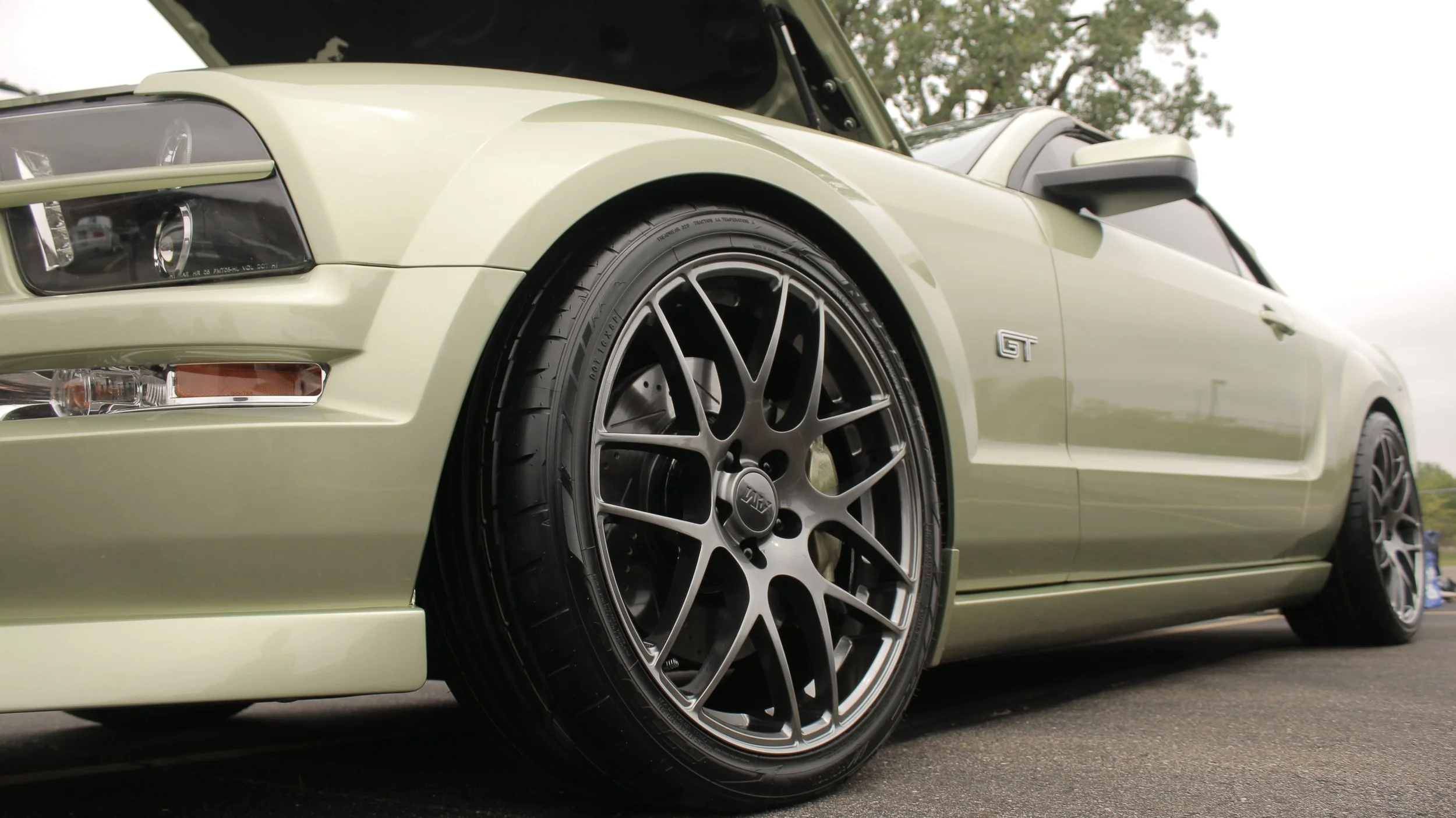 Close-up of a light green sports car showing the front left headlight, tire, wheel, and a small GT badge on the side.