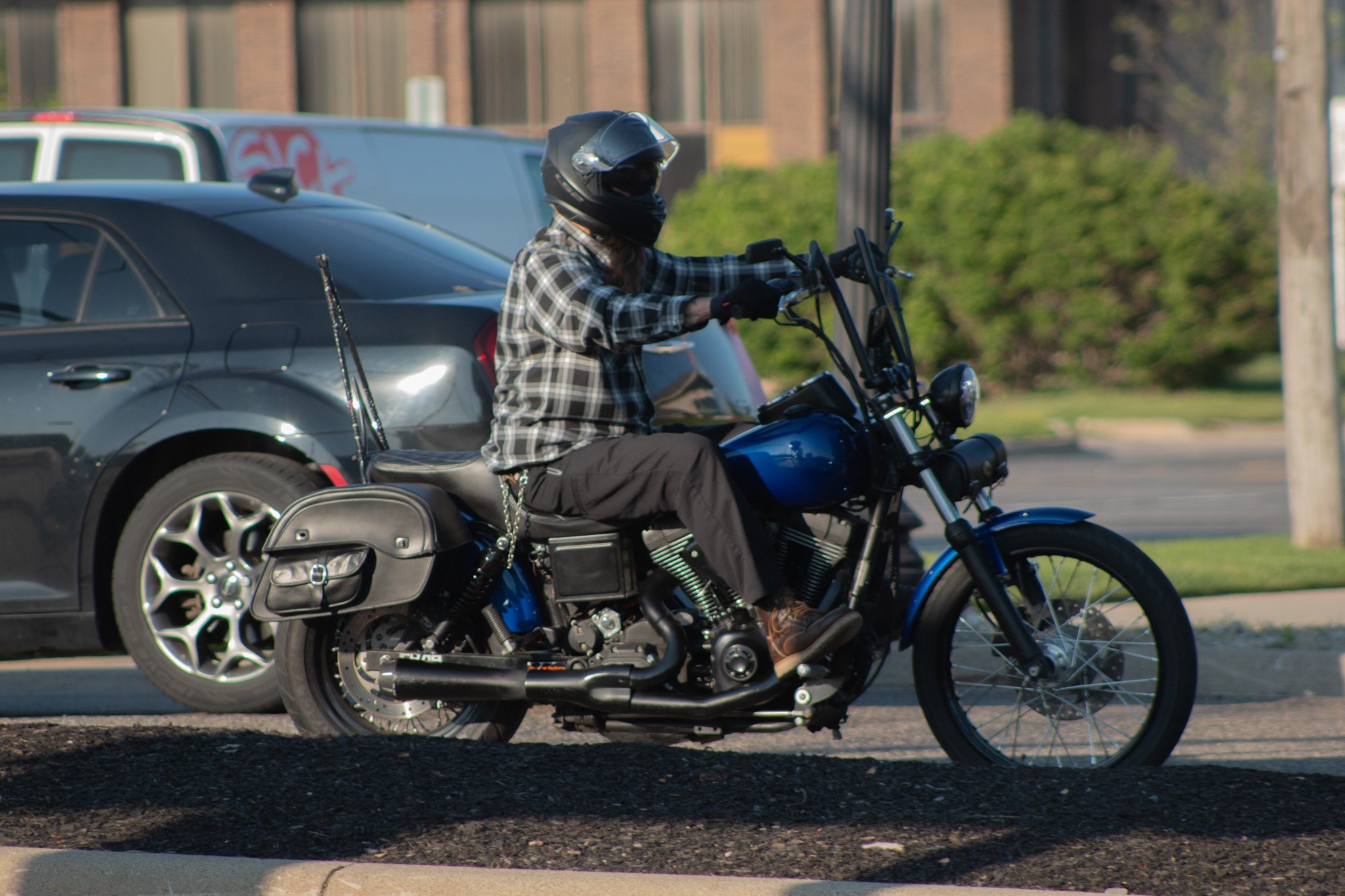 A person wearing a black helmet and plaid shirt is riding a black and blue motorcycle along a city street, with cars and trees in the background.