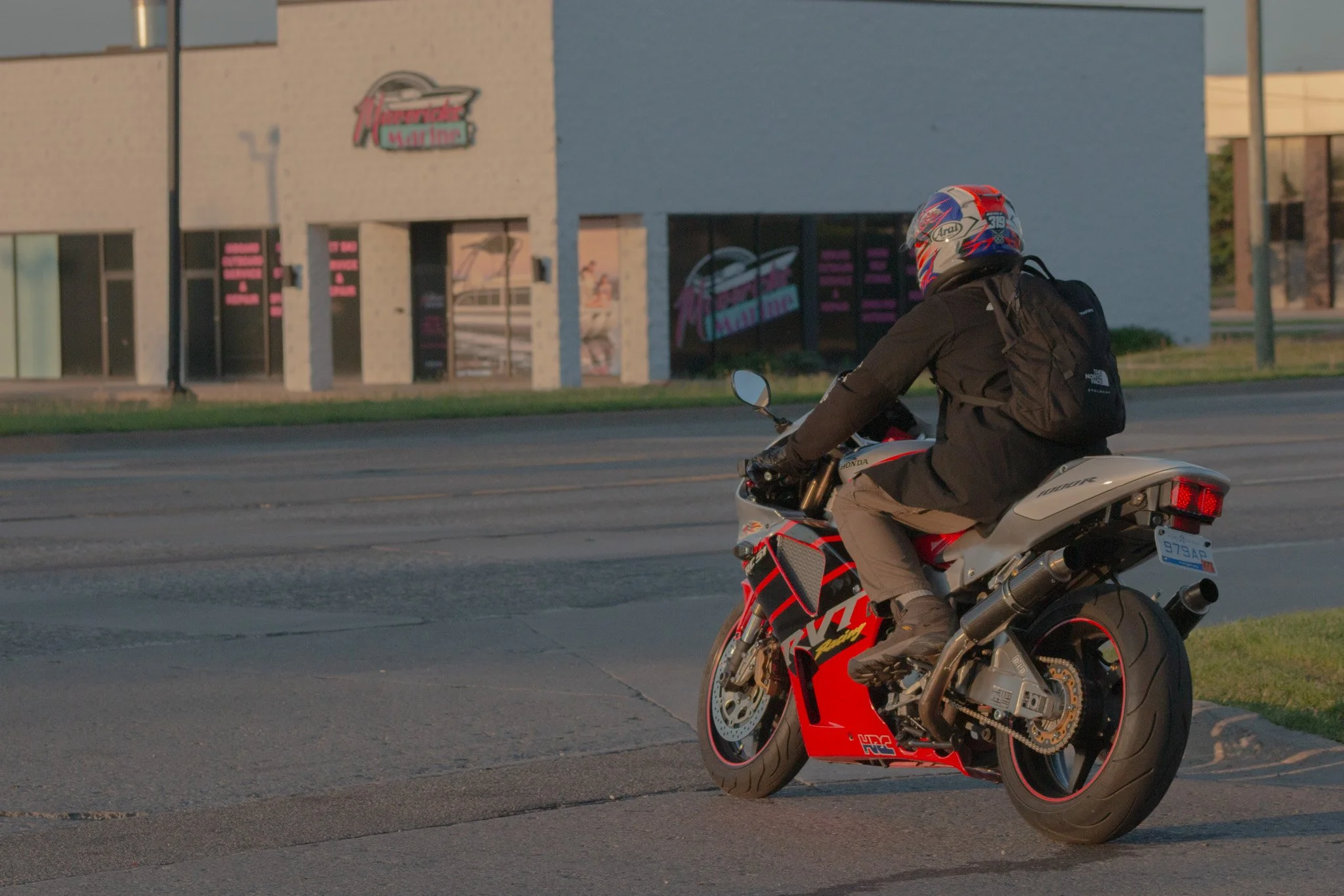 A motorcyclist wearing a blue, red, and white helmet and a black jacket rides a red and black Honda motorcycle on a city street during sunset. In the background, there are commercial buildings, including one with a sign that reads 'Martins'.