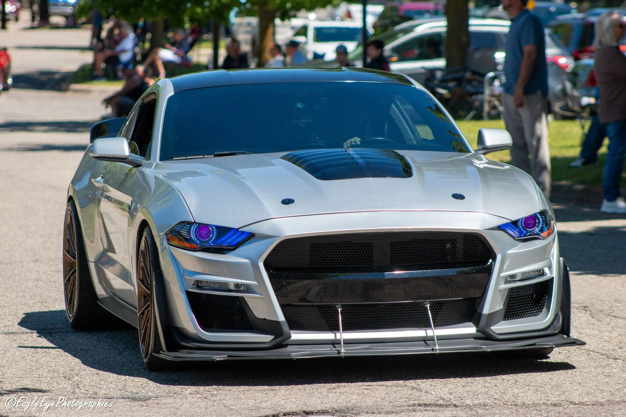 Front view of a silver modern sports car with custom headlights, parked outdoors with people sitting on benches and standing nearby, trees, and cars in the background.