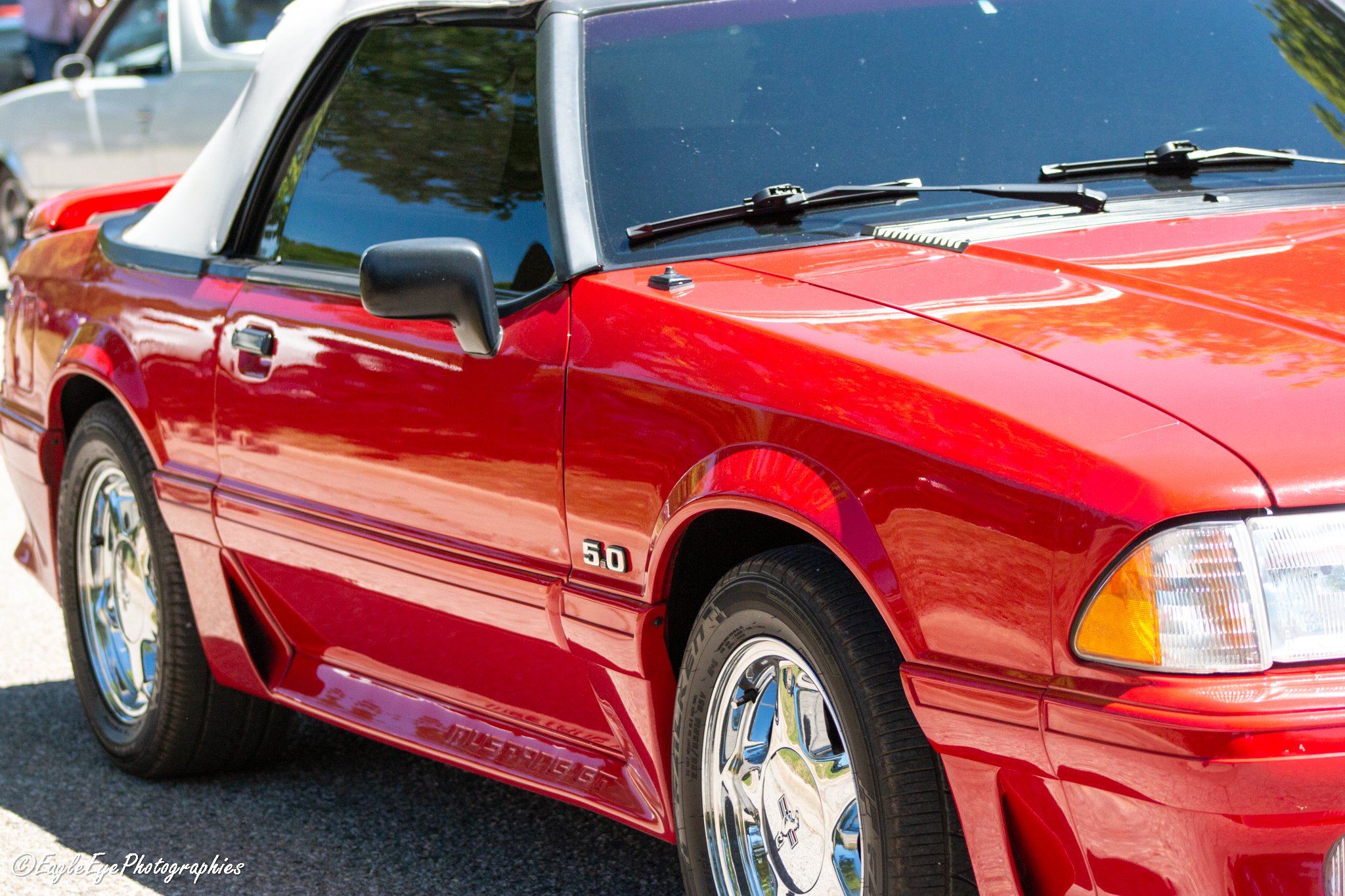 Close-up of a red vintage Ford Mustang with a 5.0 badge on the front fender, parked outdoors. Reflective chrome wheels and black side mirrors visible, with trees and sky reflected on the windshield.