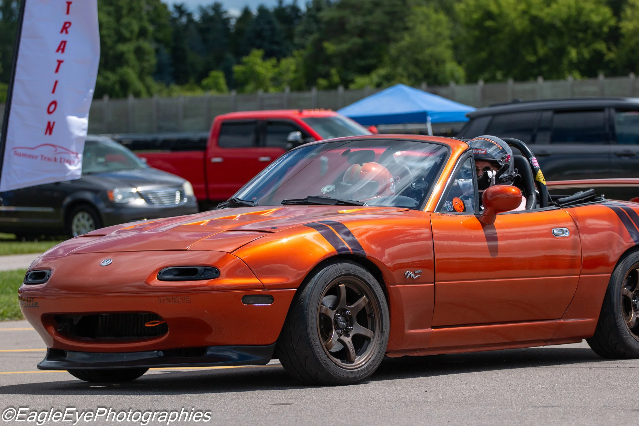 A bright orange Mazda Miata sports car with black racing stripes on the hood, driven by a person wearing a helmet, participating in a track day event at the Summer Track Days, with a red truck, black car, and a blue canopy in the background.