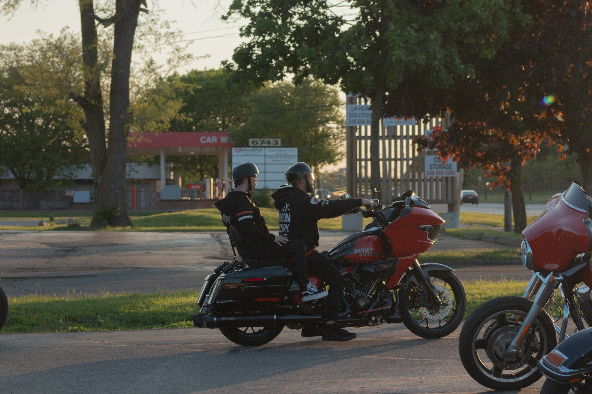 Two motorcyclists sit on a Harley Davidson motorcycle parked on the side of the road, with trees and signs in the background during sunset.