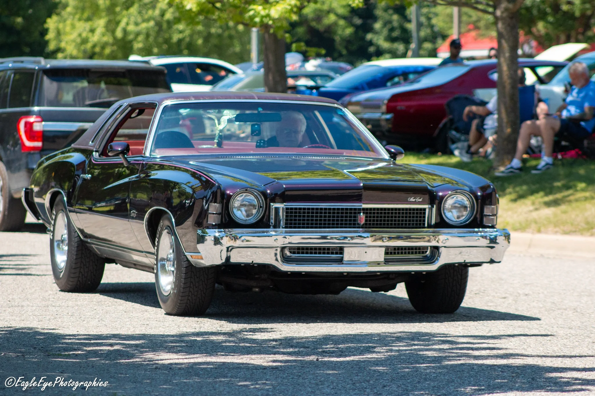 A shiny black vintage Chevrolet Corvette at a car show, parked outdoors on a sunny day, with other cars and people in the background.