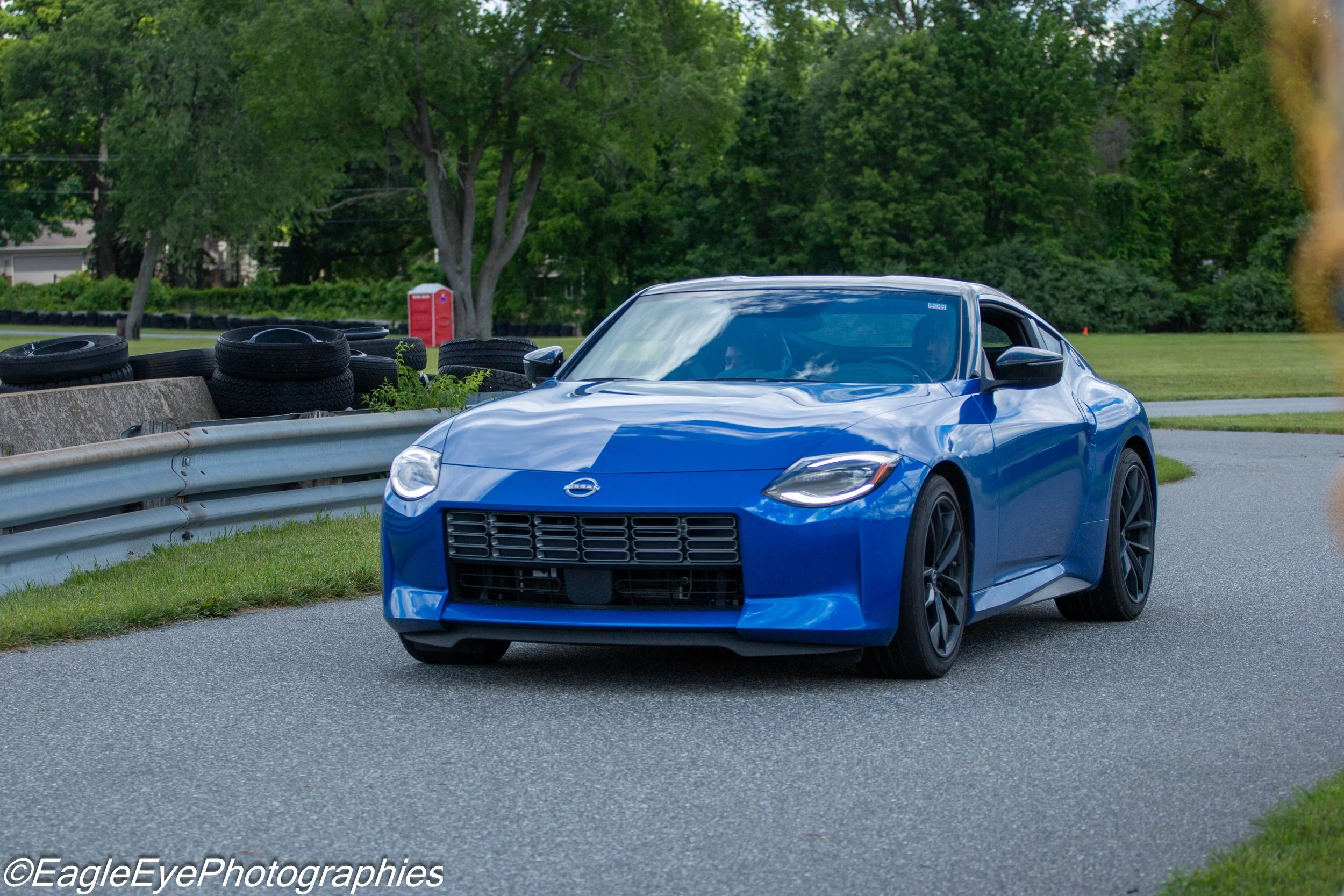 A blue sports car parked on the side of a road with green trees and tires stacked on a concrete barrier nearby.