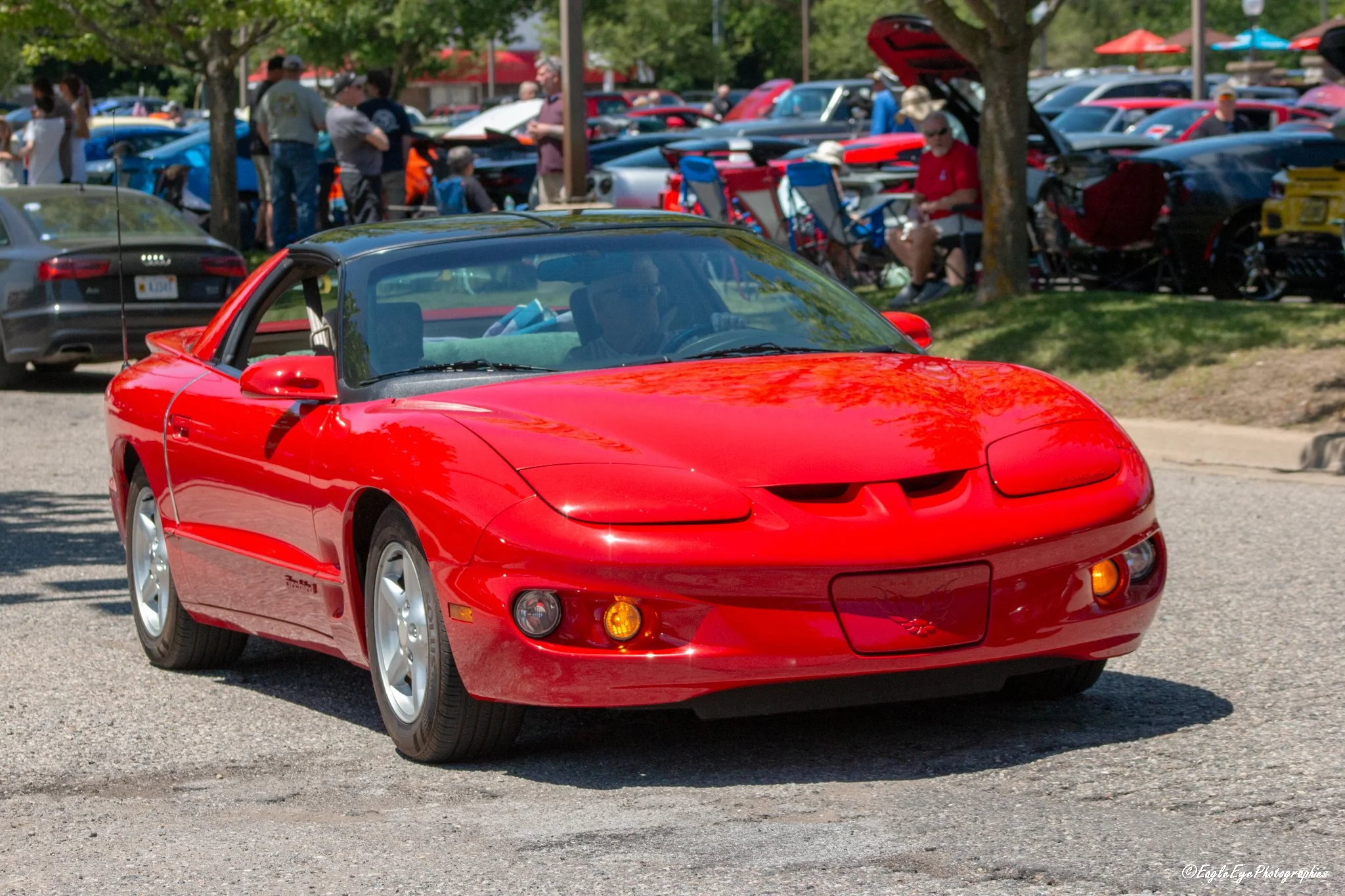 Red Chevrolet Corvette sports car parked at a car show during daytime, with multiple classic and modern cars and people in the background.