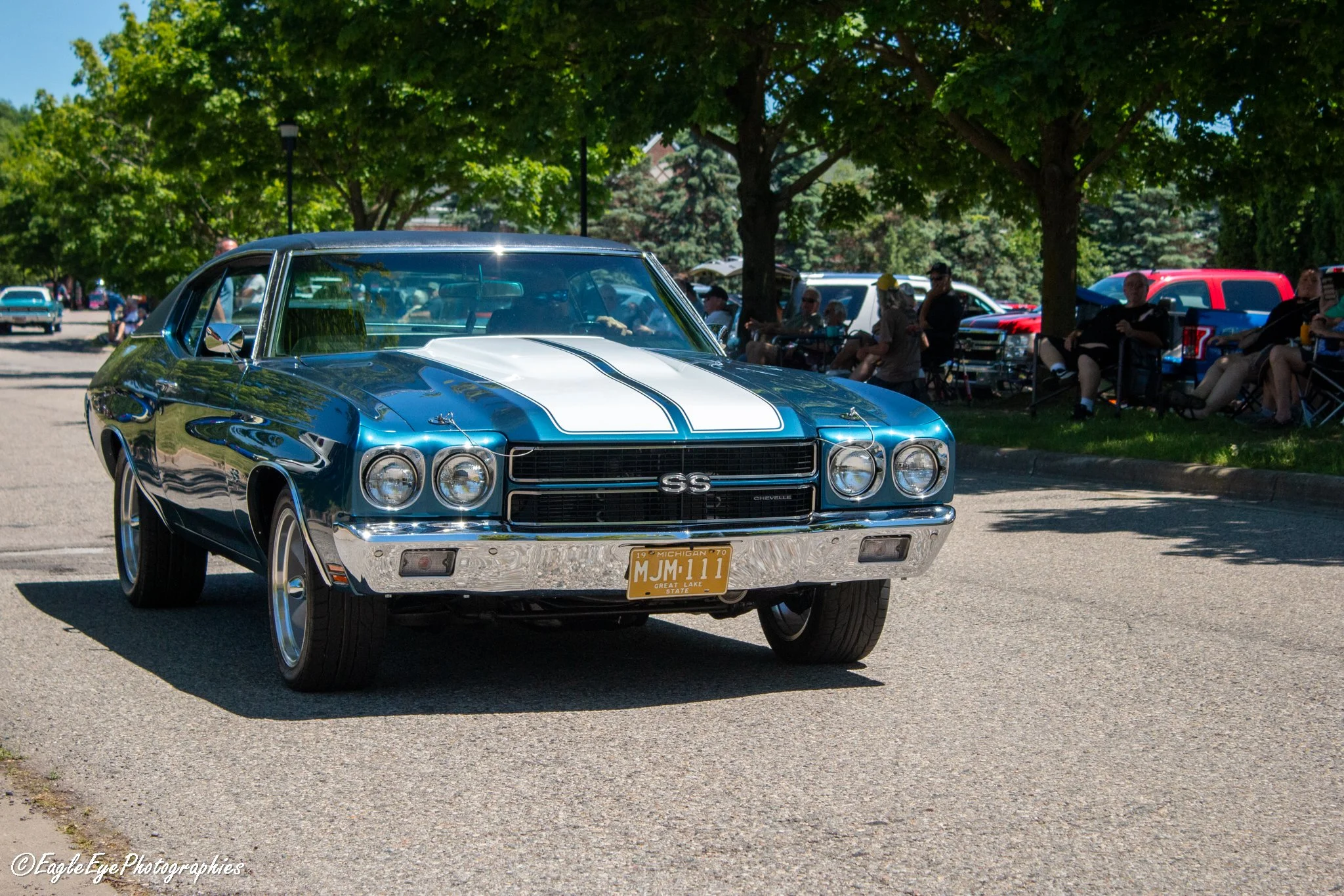A vintage blue Chevrolet Chevelle with white racing stripes on the hood and Michigan license plate, parked at a classic car show with people sitting under trees in the background.