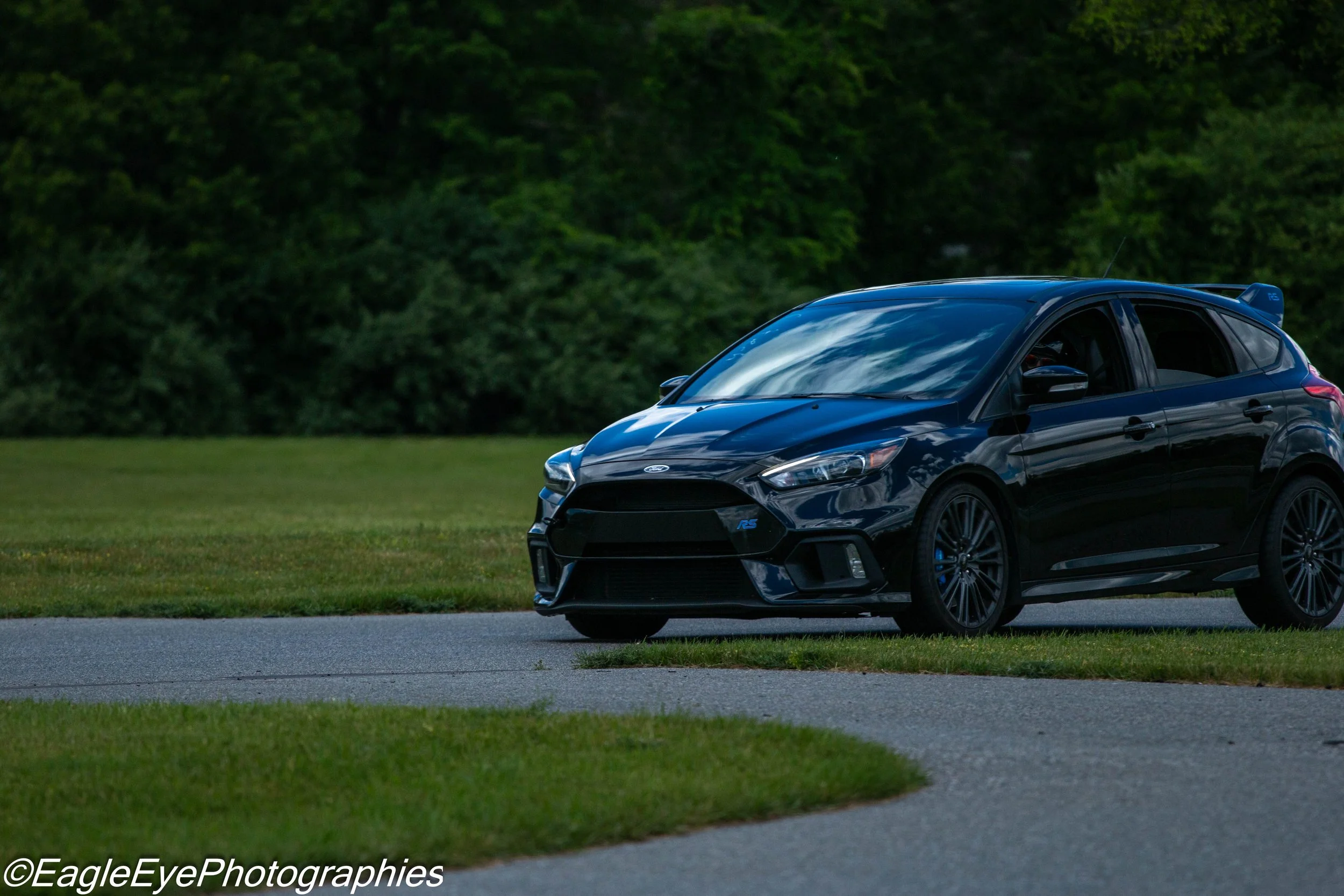 A black Ford Focus RS parked on a grassy roadside with a background of green trees, during daylight.