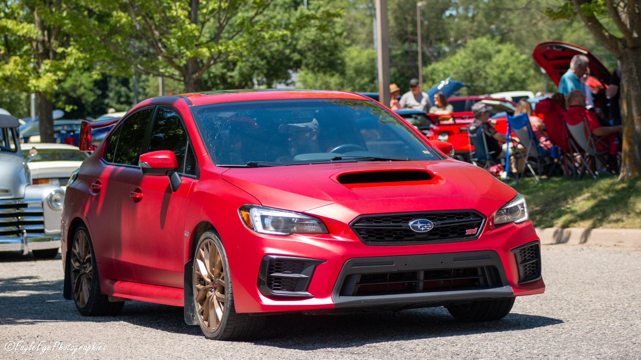 A red Subaru WRX STI car parked outdoors at a gathering with people sitting on chairs under trees in the background.