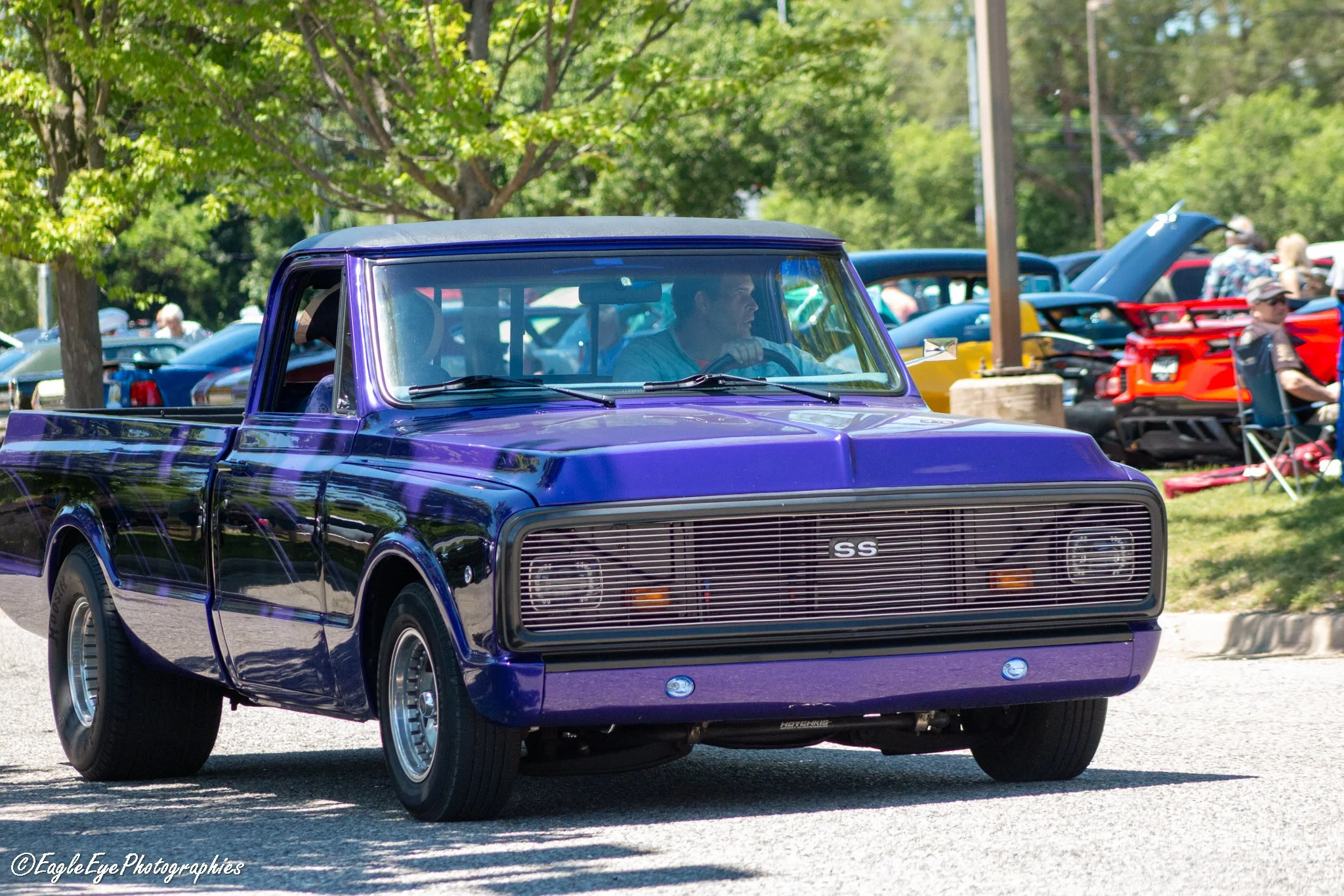 A vintage purple pickup truck driving on a street during a car show event, with trees and people in the background.