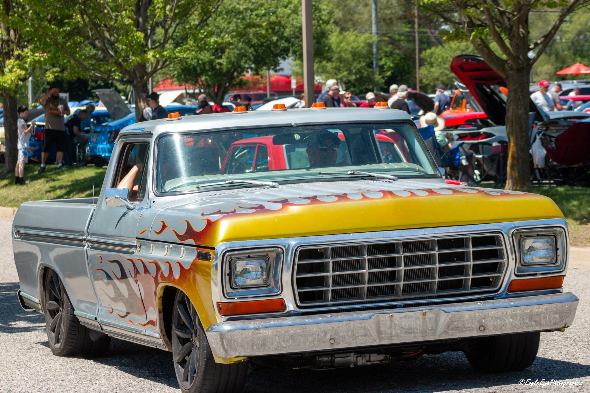 A vintage pickup truck with flame decals parked at a car show, surrounded by people and other classic cars under trees.