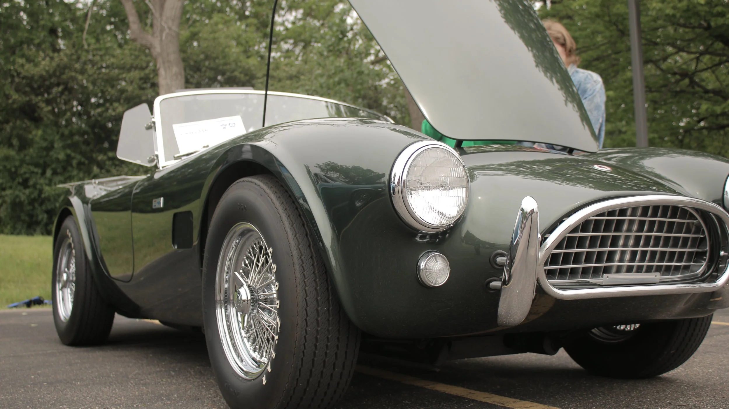 Close-up of a vintage black convertible sports car with a raised hood, chrome wheel spokes, and a front grille, parked on pavement with green trees in the background.