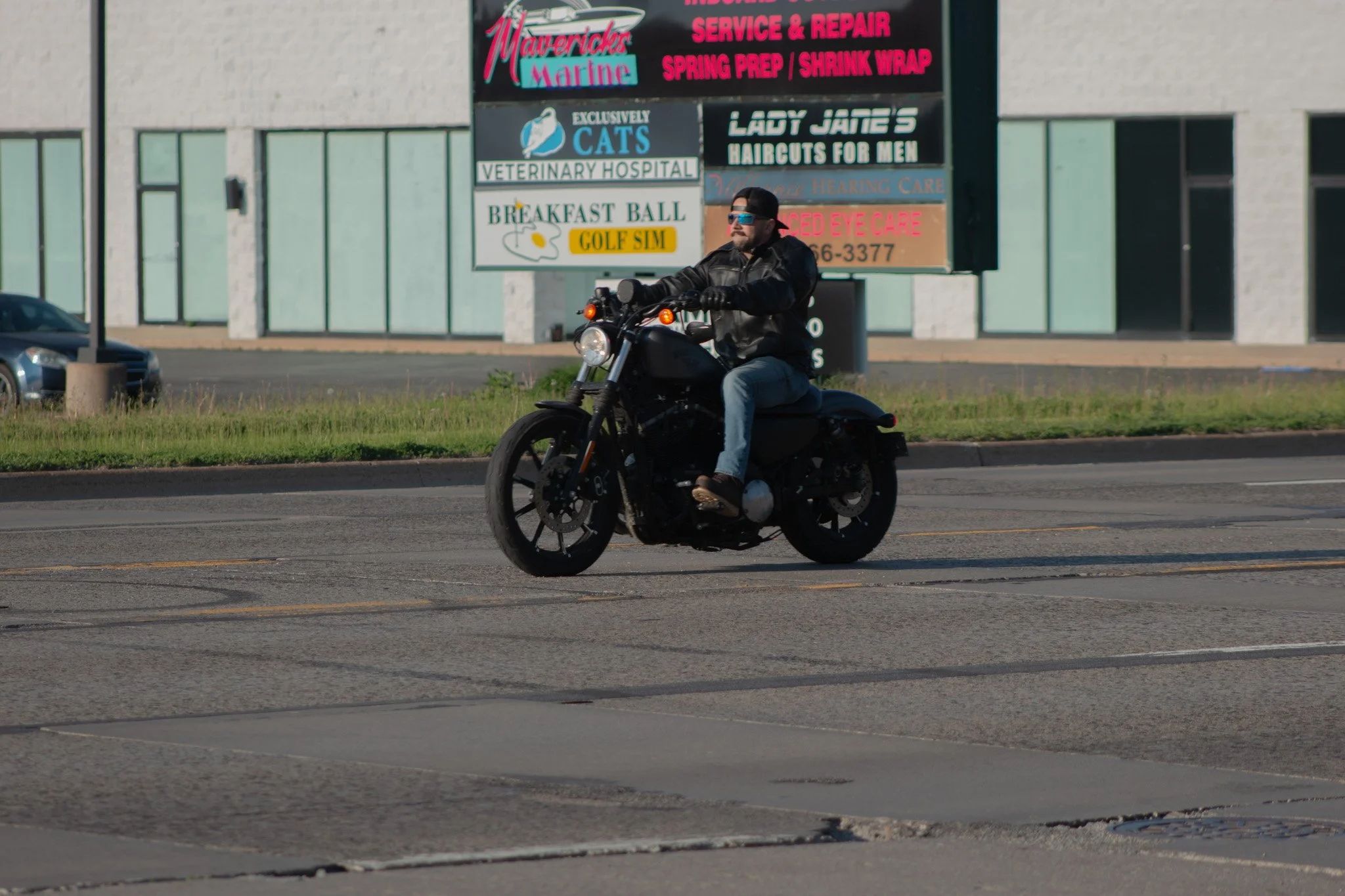 A man riding a black motorcycle on a city street, wearing a black leather jacket, jeans, sunglasses, and a black cap backwards, with a retail strip mall in the background.