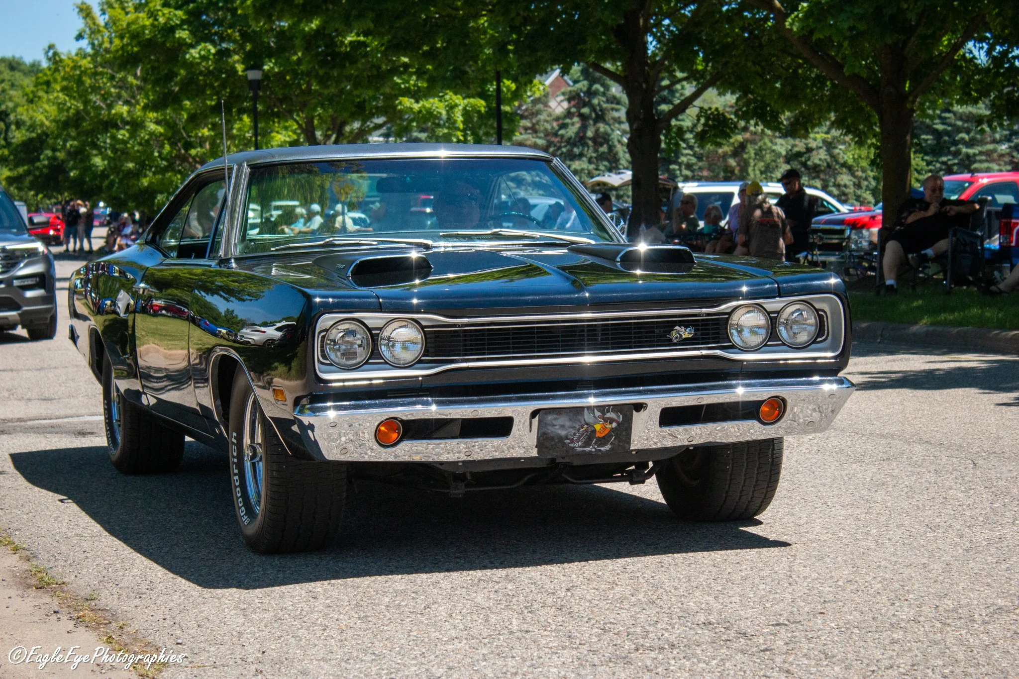 A black vintage muscle car with a sleek design, dual hood air scoops, and chrome front bumper parked on a paved surface at an outdoor car show or gathering. In the background, there are green trees, other cars, and people sitting and walking.