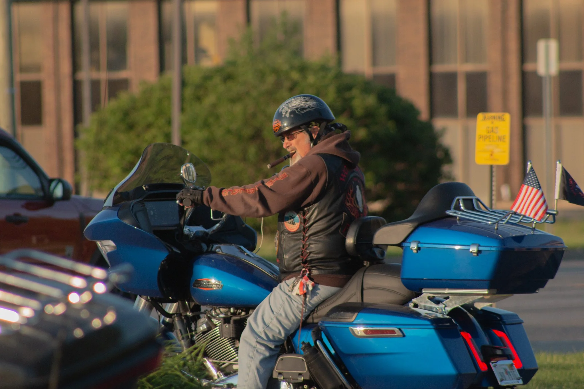 A man riding a blue motorcycle with a windshield, wearing a black helmet with a Harley Davidson logo and sunglasses. The motorcycle is decorated with small American flags, and there is a yellow sign in the background that reads 'Gas Pipeline.'