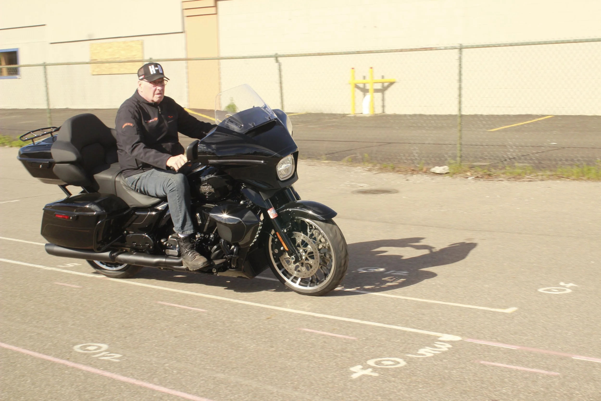 A man riding a black Harley-Davidson touring motorcycle in a parking lot, wearing a black jacket and cap, with a chain lock on the front wheel, and a large backrest on the motorcycle, near a chain-link fence and a beige building.