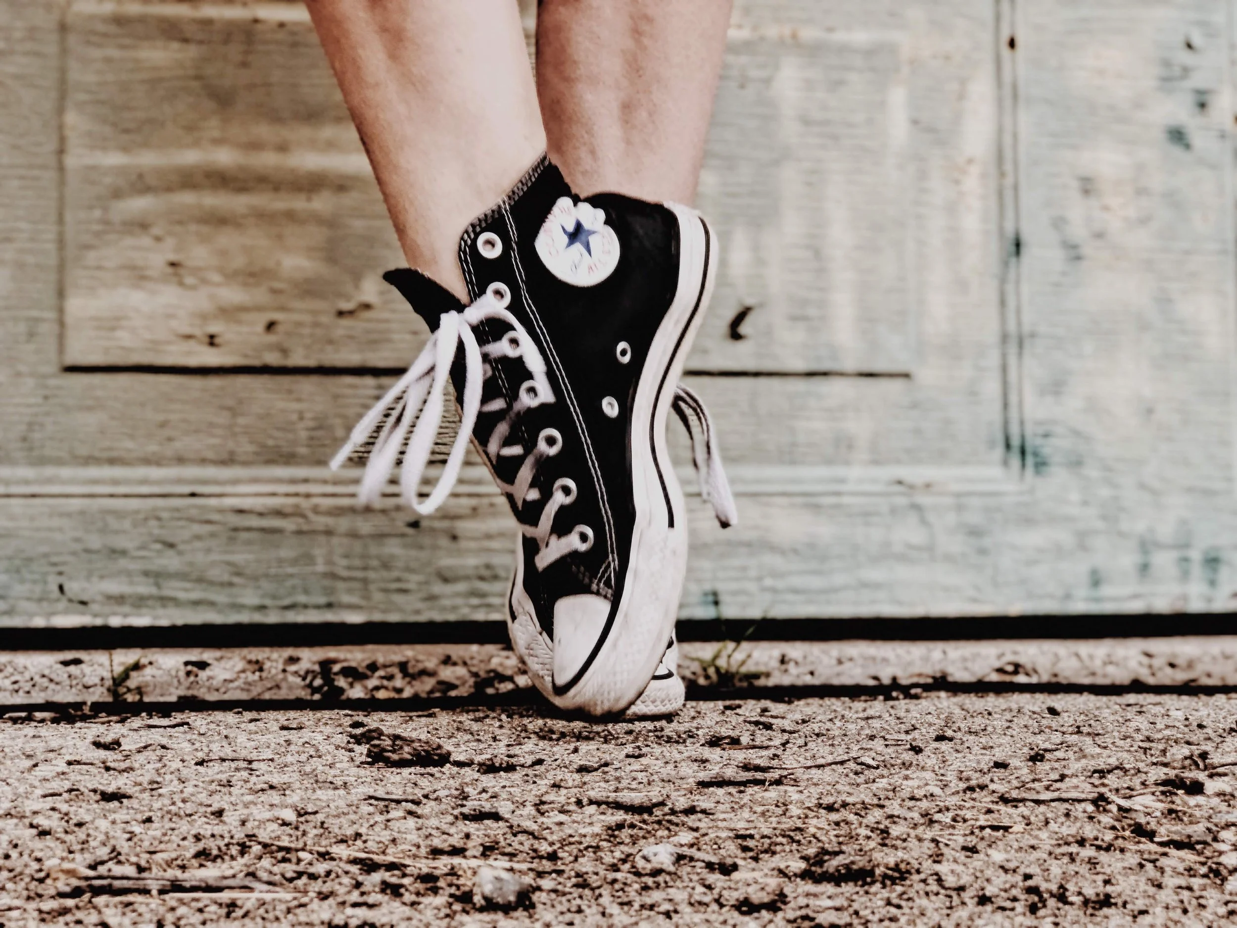 Close-up of a person wearing black high-top Converse sneakers standing on a wooden deck with small rocks or gravel underneath.