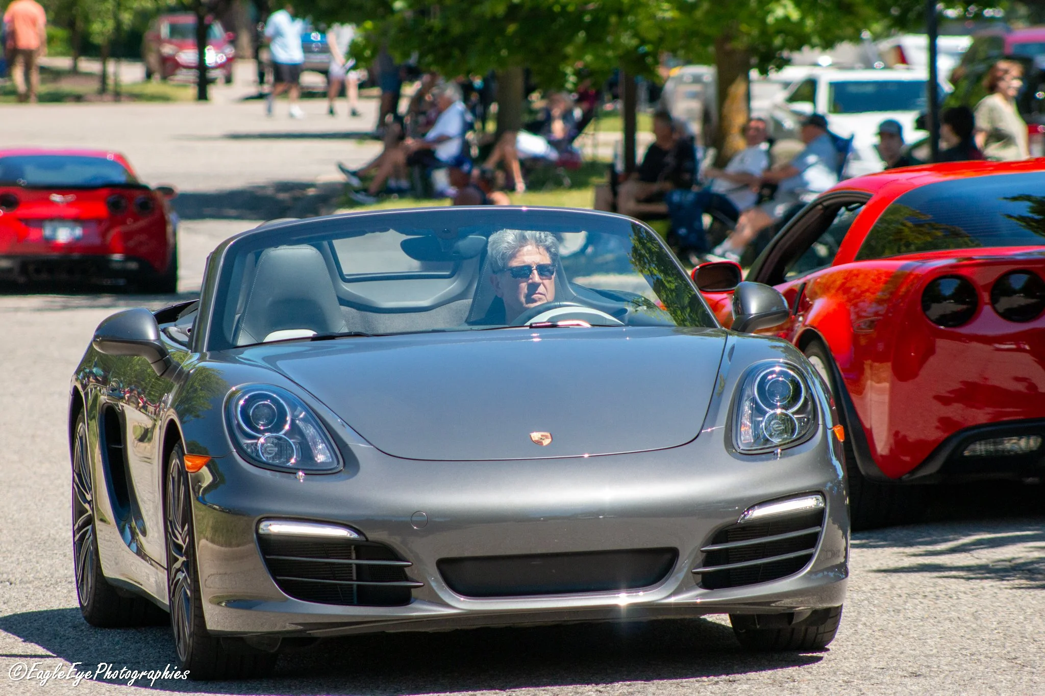 A silver Porsche convertible car with a man wearing sunglasses inside, parked in a lot with other cars and people sitting under trees in the background.