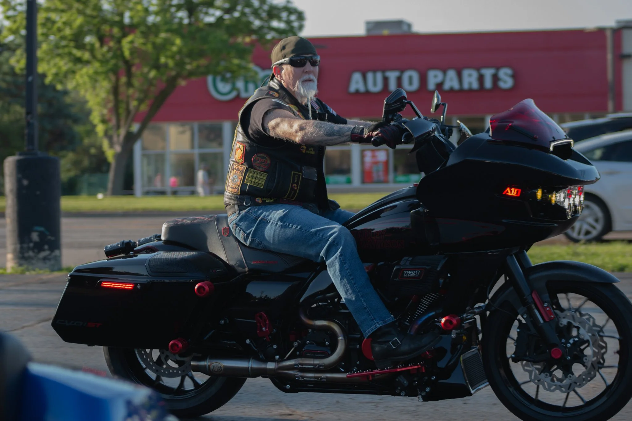 An older man riding a black motorcycle on a street with a red auto parts store in the background. He is wearing sunglasses, a black cap, a black vest with patches, and blue jeans.