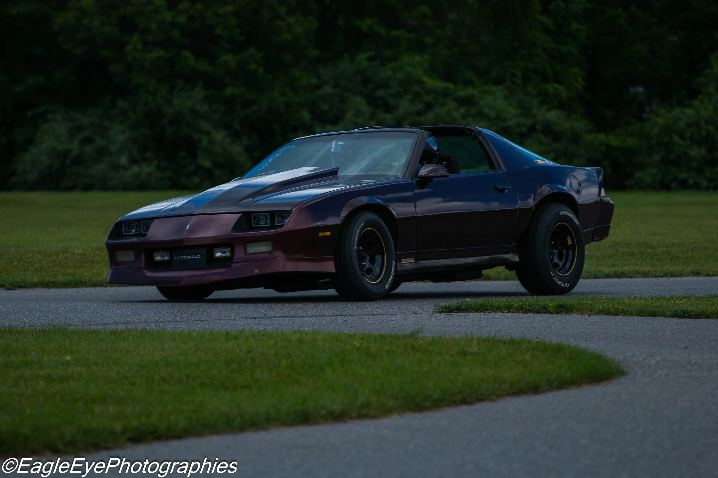A classic two-tone black and dark red Chevrolet Camaro sports car parked on a grassy area with a dense tree line in the background.