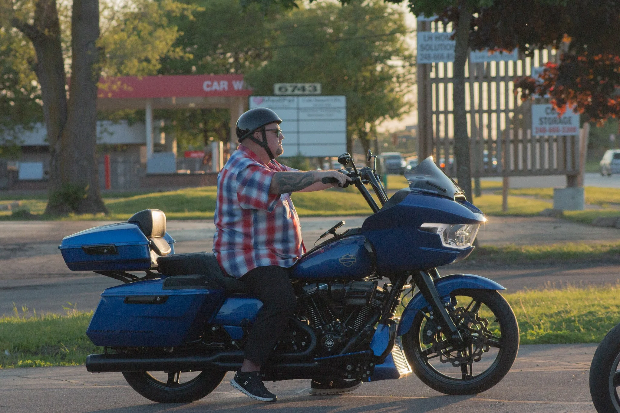 A man wearing a black helmet, sunglasses, and a red plaid shirt riding a blue Harley-Davidson motorcycle on a street during sunset. There are trees and signs in the background.