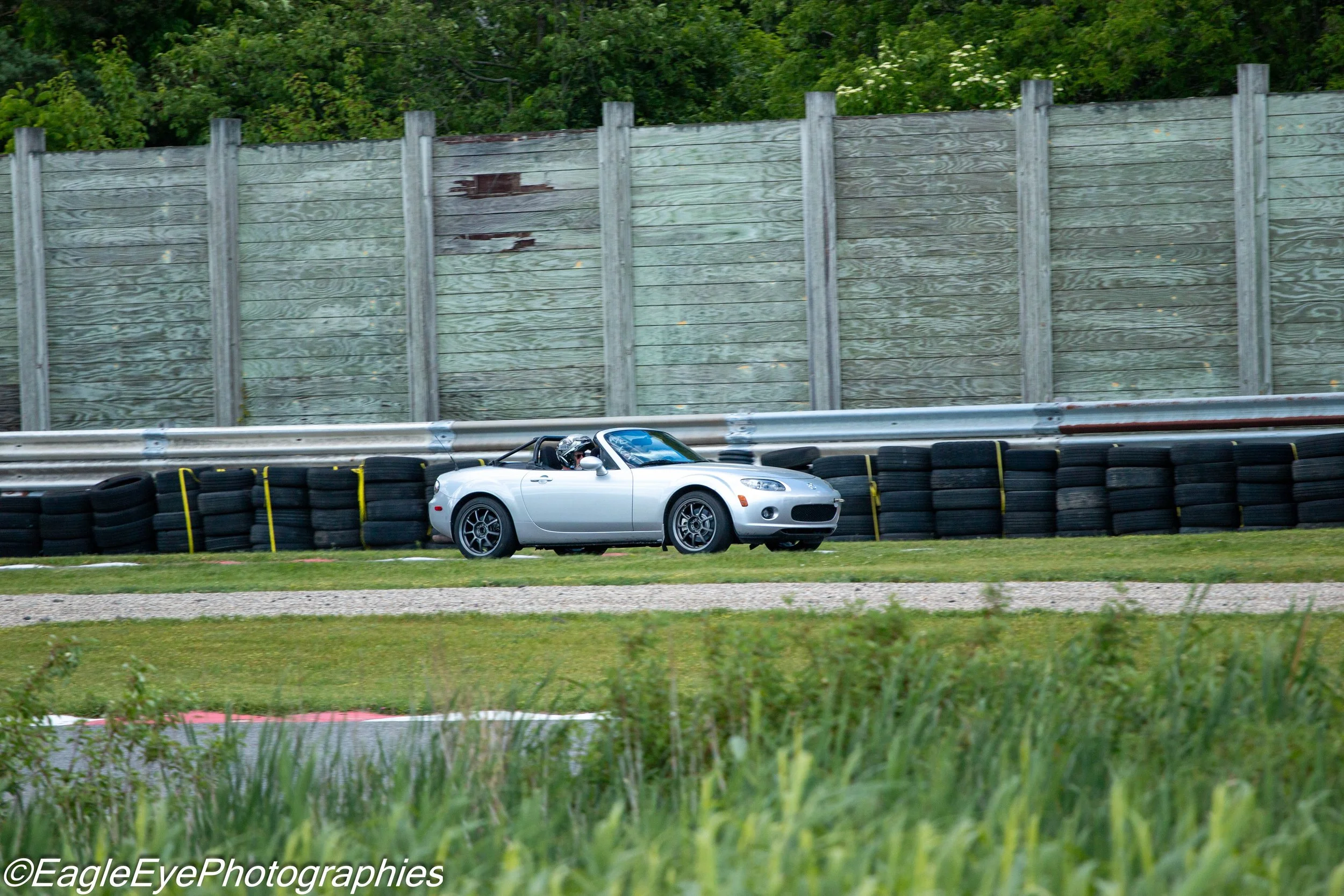 A silver sports car drives along a race track, with a green wooded area in the background and a tall weathered wooden fence separating the track from the forest. Tire barriers are visible at the side of the track.