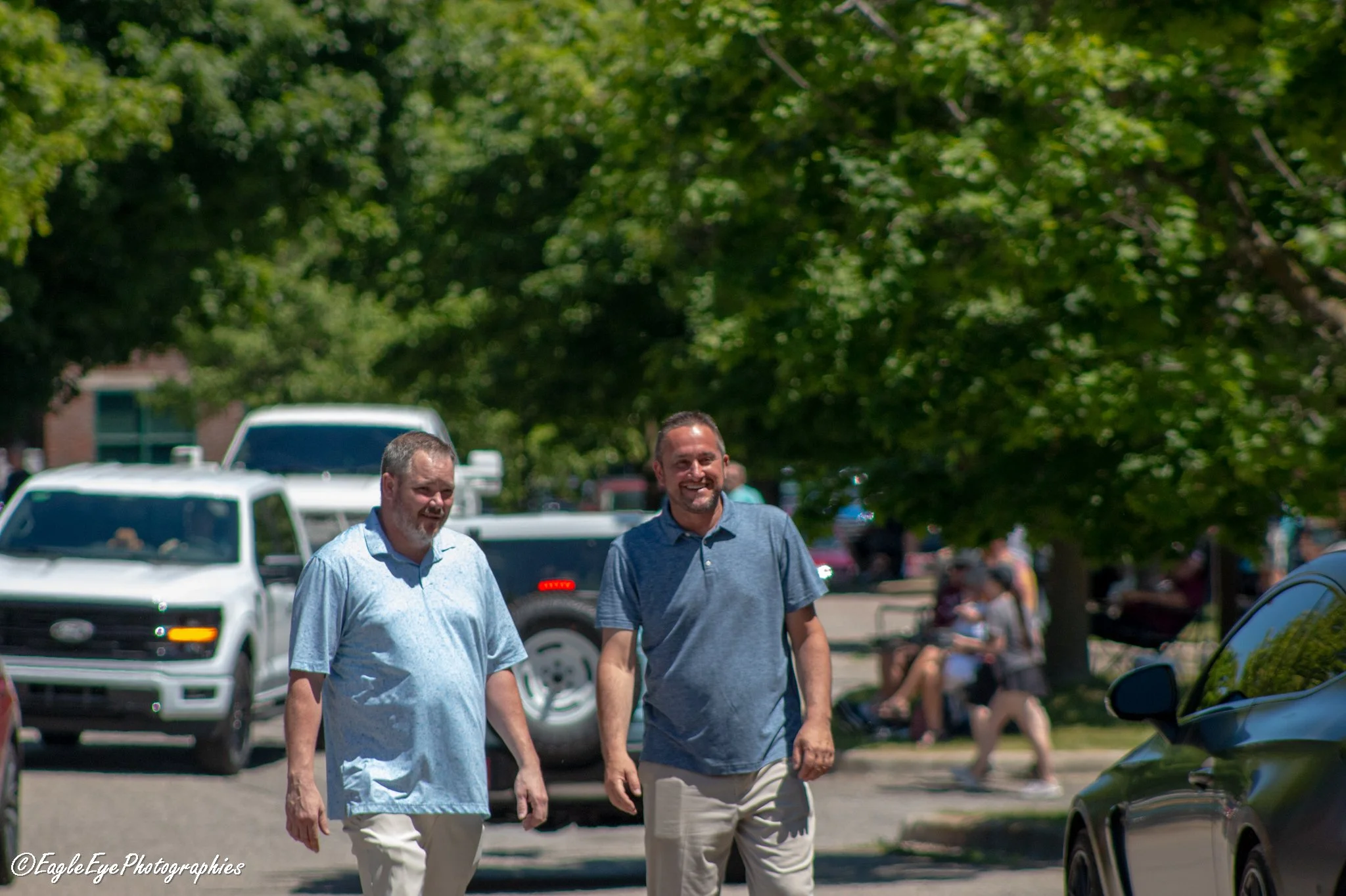 Two men walking outdoors on a sunny day with trees and cars in the background.
