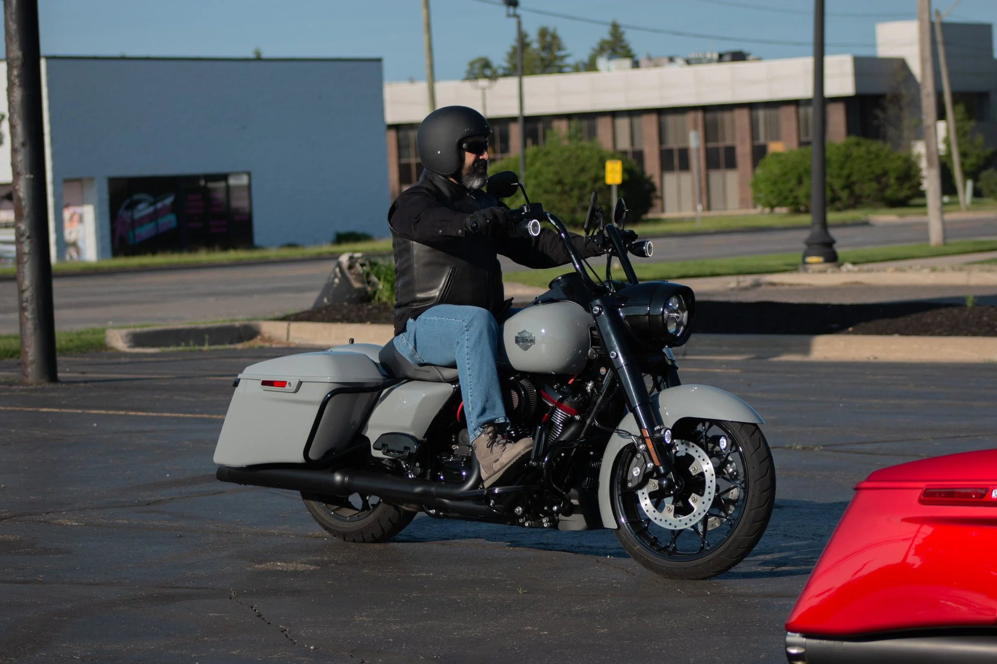 A man wearing a black helmet, sunglasses, a black jacket, and jeans riding a white motorcycle in a parking lot. There are cars and a building in the background.