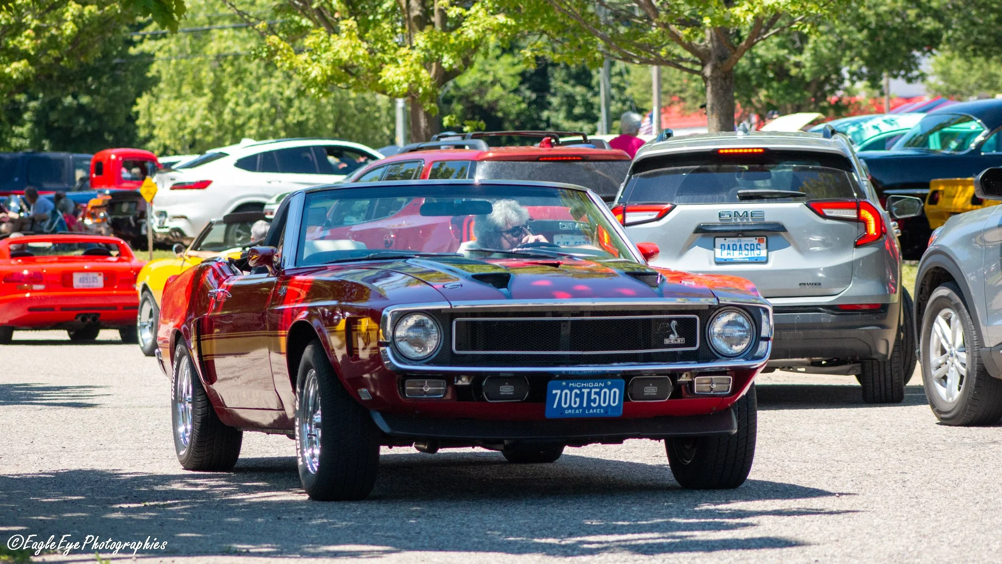 Red vintage Shelby Mustang car with Michigan license plate driving through a crowded parking lot filled with various modern cars, trees in the background, and people sitting in the shade.