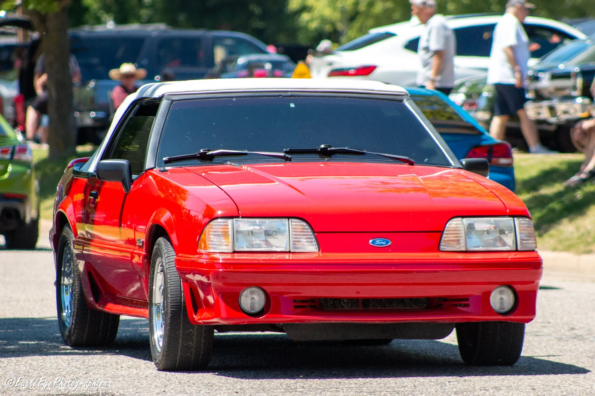 A red vintage Ford Mustang car with black-tinted windows parked outdoors at a car show with other cars and attendees in the background.