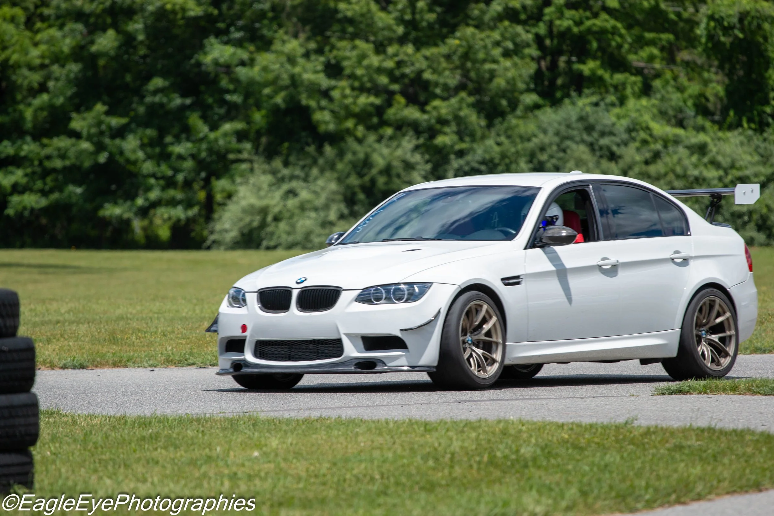 A white BMW race car on a grassy field with a green trees background, with a rear wing and racing modifications.