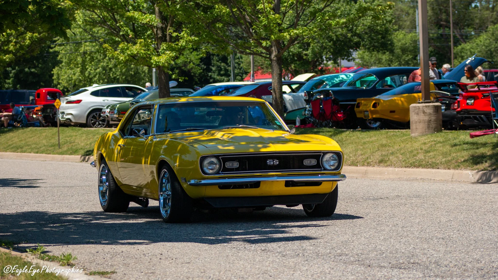 A yellow vintage Chevrolet Camaro SS car driving on a street during a car show, with other classic and modern cars parked in the background under trees.