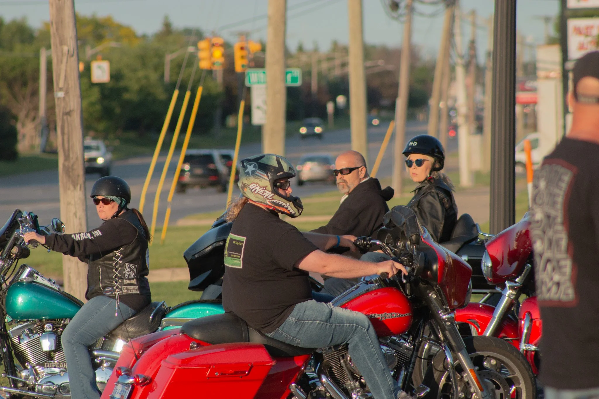 Multiple motorcyclists, some wearing helmets and sunglasses, are riding on a road with cars and trees in the background.