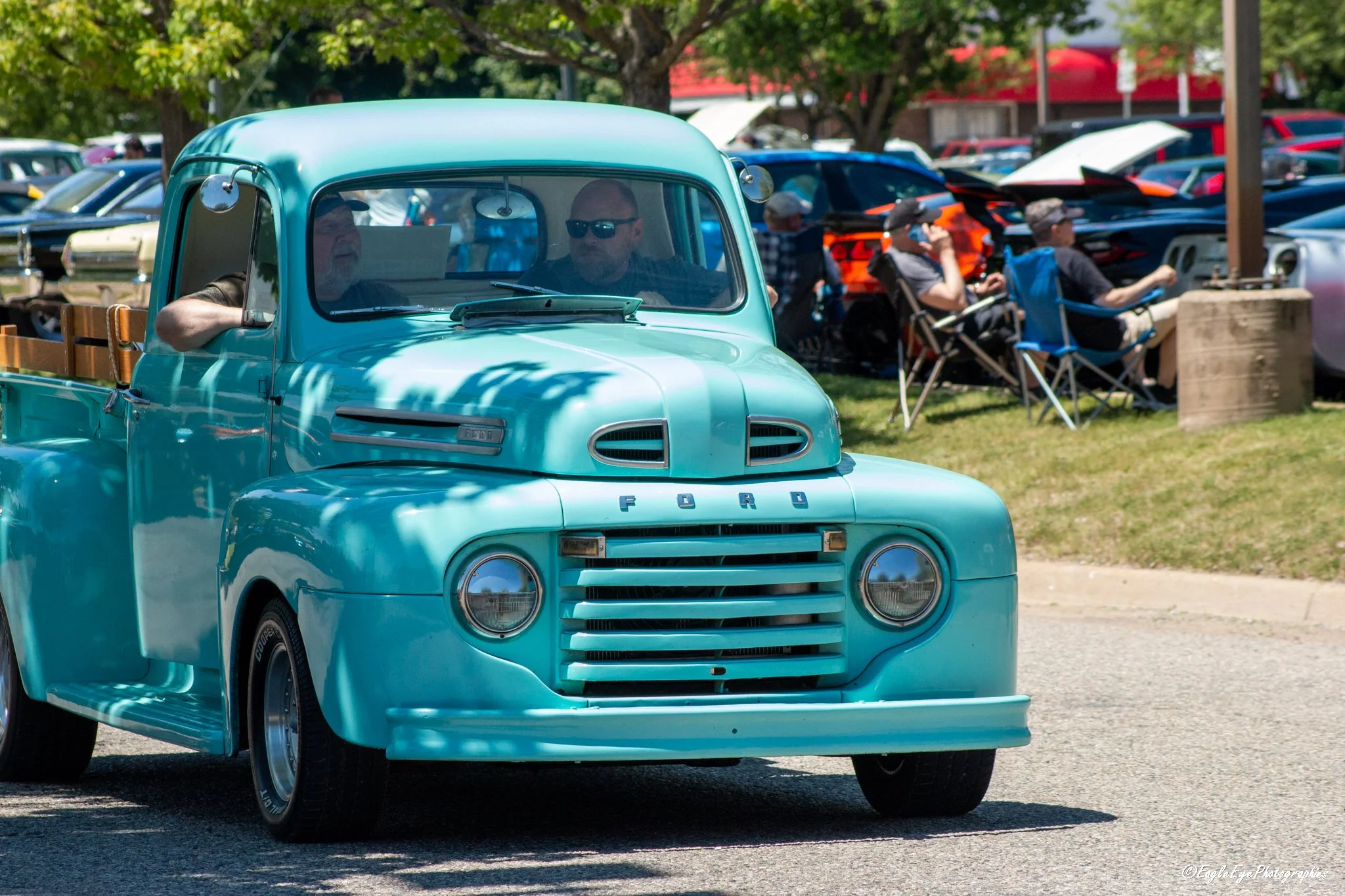 A vintage turquoise pickup truck parked outdoors at a classic car show, with people sitting in chairs and other cars visible in the background.