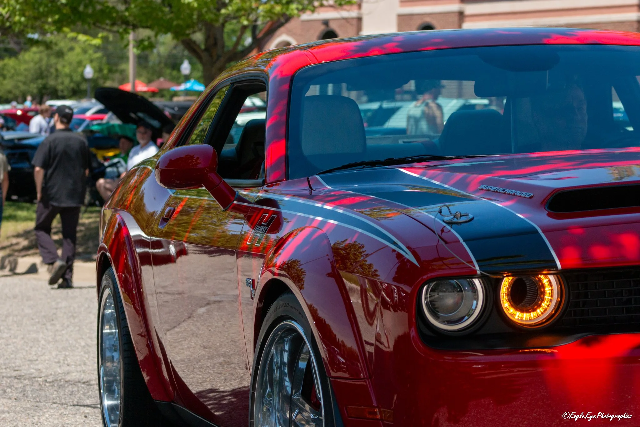 Close-up of a red sports car with racing decals, parked outdoors at a car show with people and trees in the background.
