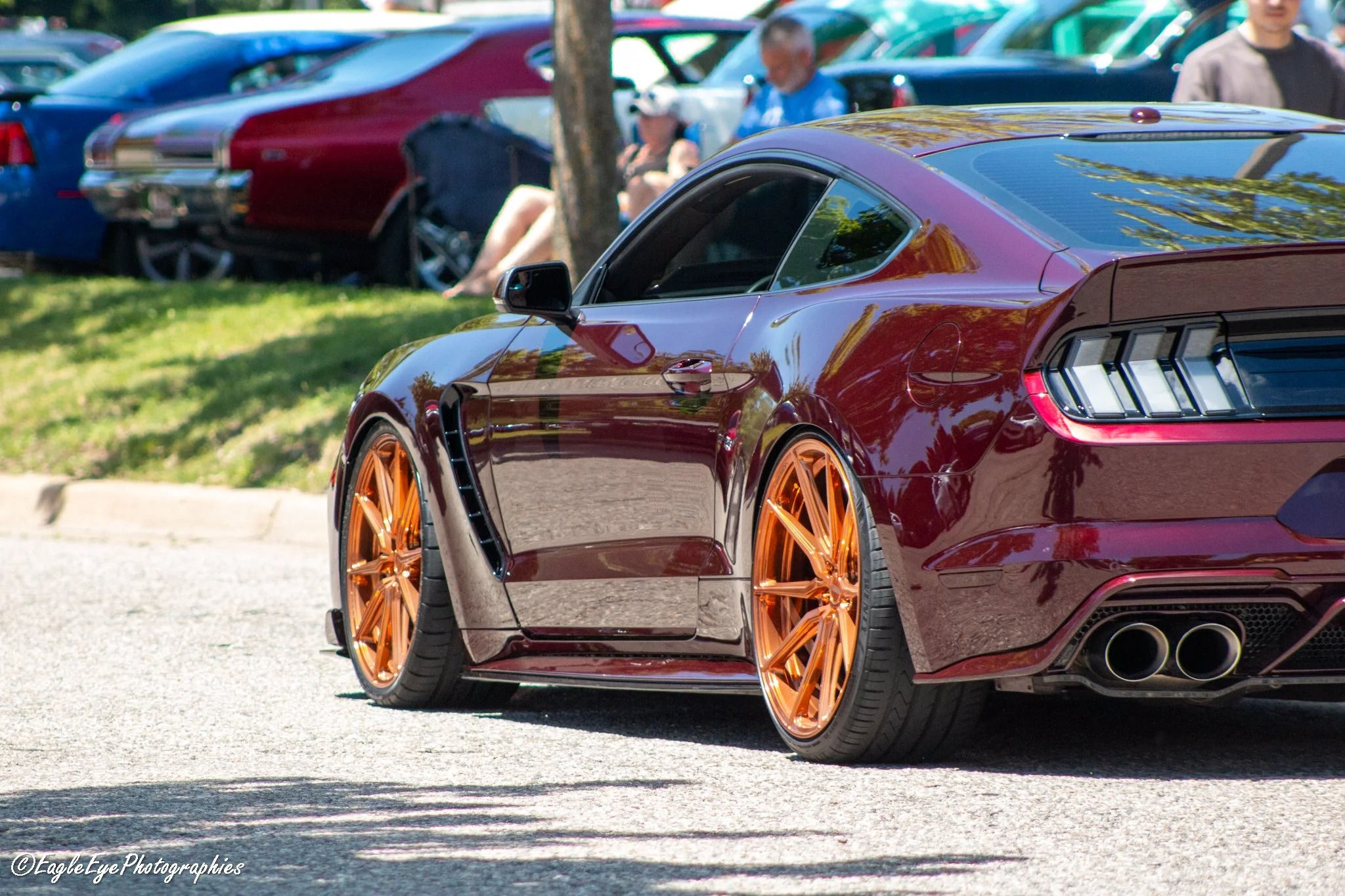Close-up of a shiny burgundy sports car with large, orange rims parked at a car show, with a background of other parked cars and people sitting and standing outdoors.