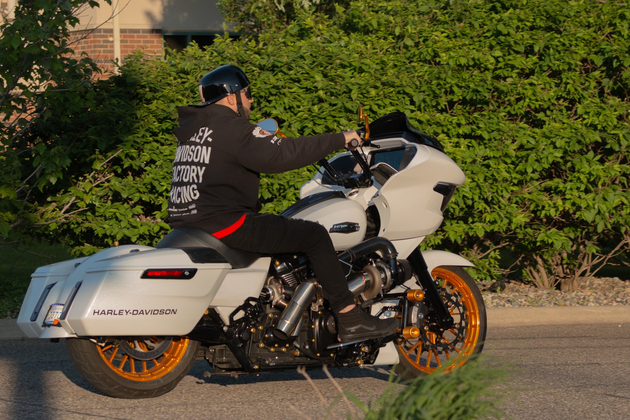 A man wearing a black Harley-Davidson hoodie, black helmet, and sunglasses riding a white Harley-Davidson touring motorcycle with orange wheels on a street in front of green bushes and a building.