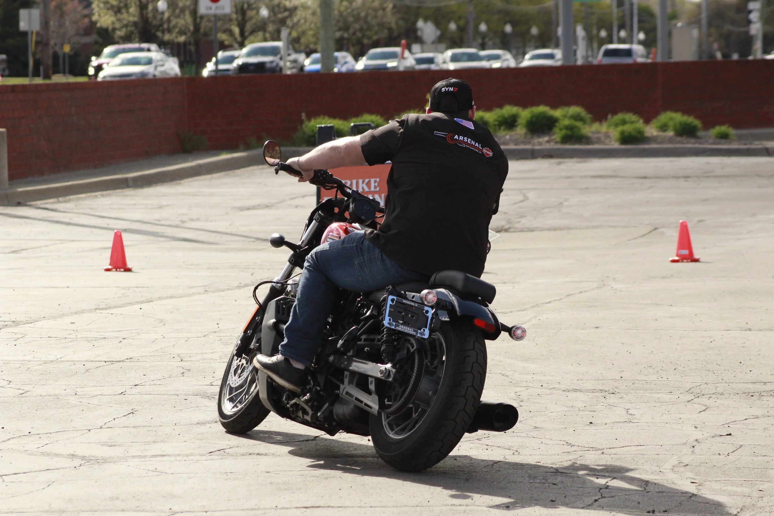 A man riding a black motorcycle around orange traffic cones in an open lot. The man is wearing a black shirt with logos and a black cap, with parked cars and a brick wall in the background.