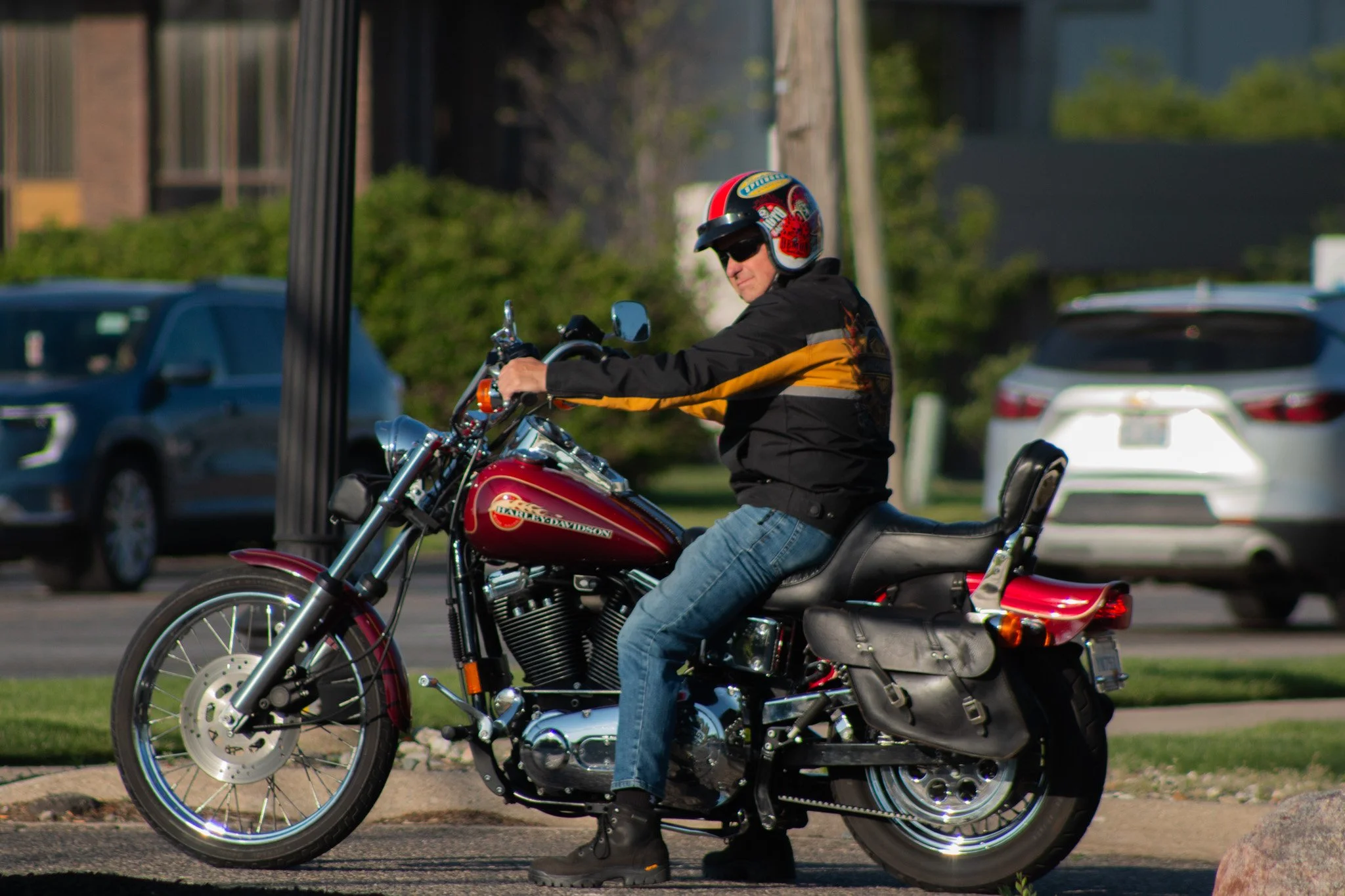 A man wearing a colorful helmet, sunglasses, and a black and orange jacket riding a red Harley-Davidson motorcycle on a street with parked cars and greenery in the background.