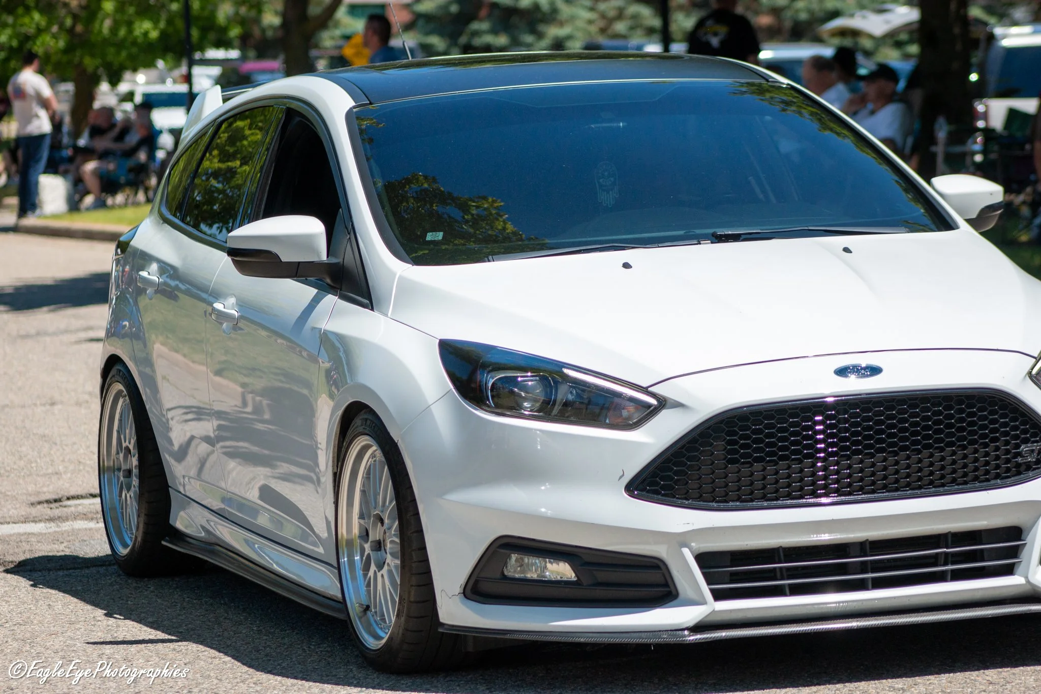 A white hatchback car with black accents parked on a street in a park-like setting during daytime, with trees, people, and other vehicles in the background.