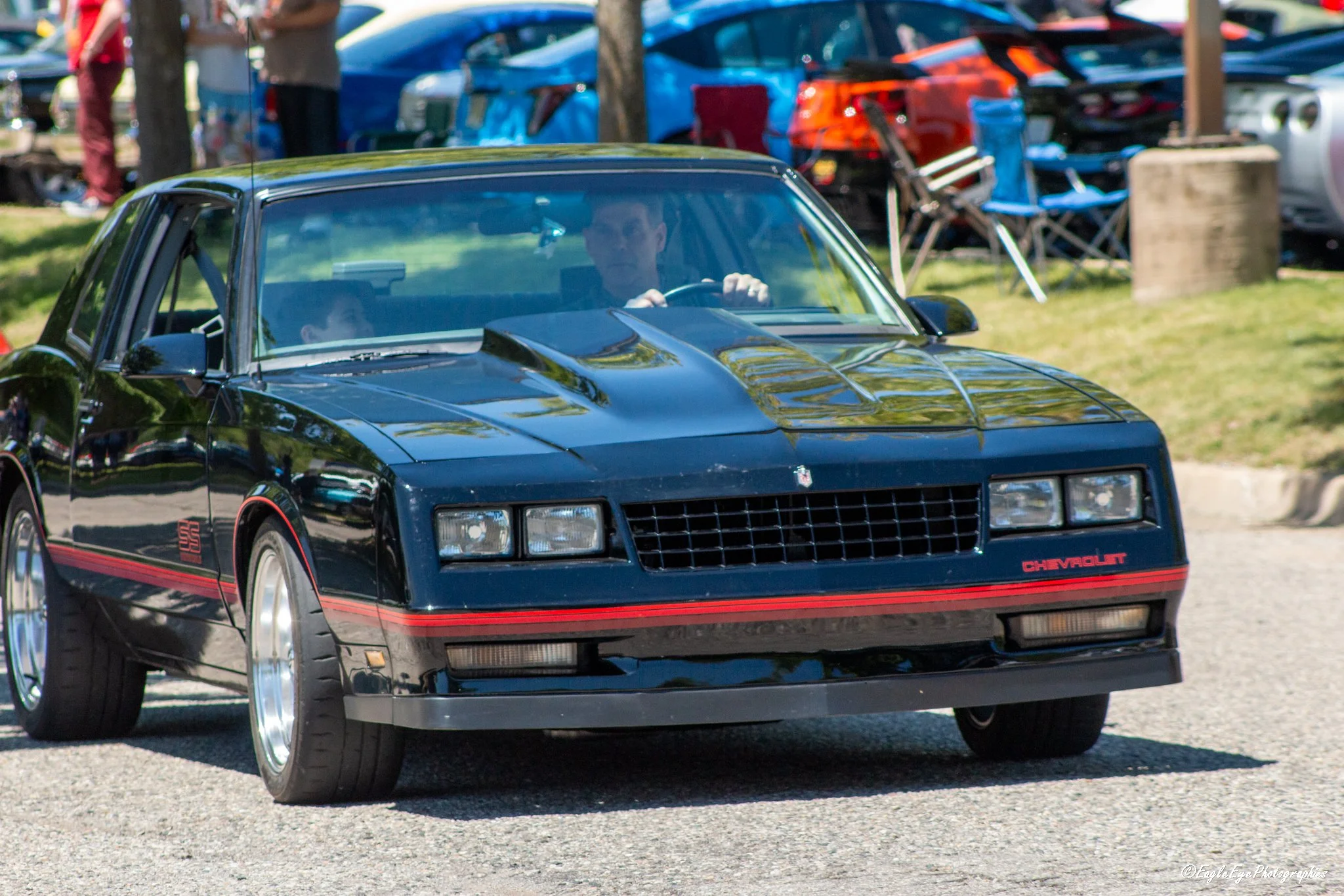 A black Chevrolet sports car with red accents and a glossy finish parked outdoors at a car show. The car has a sleek design, with a person driving and a young boy in the passenger seat. Background includes other cars, trees, and people.