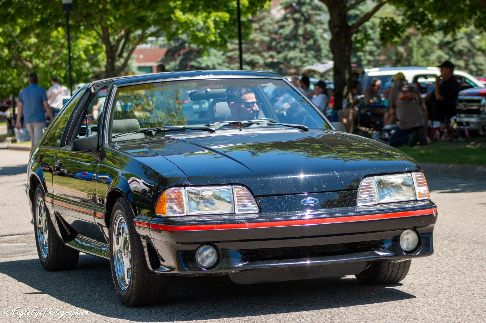 A black vintage Ford Mustang car at a car show or event with trees and people in the background.