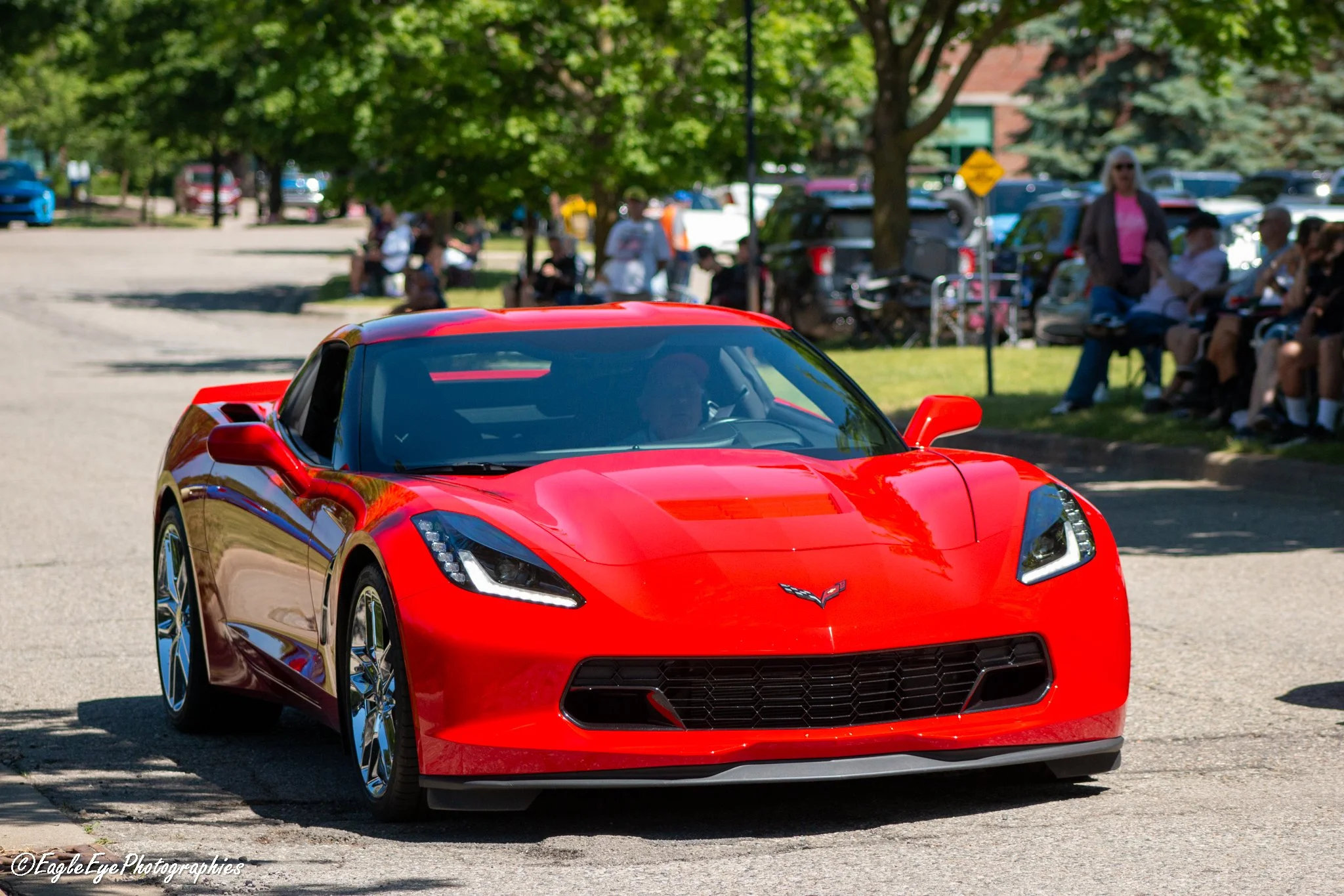 Close-up of a red sports car parked in a lot with people sitting on benches and trees in the background.