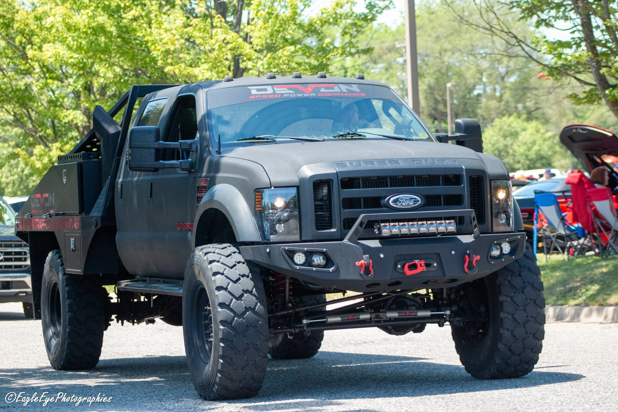 A black Ford Super Duty truck with modified rugged tires and off-road accessories, parked outdoors at a car show or event with green trees and other vehicles in the background.