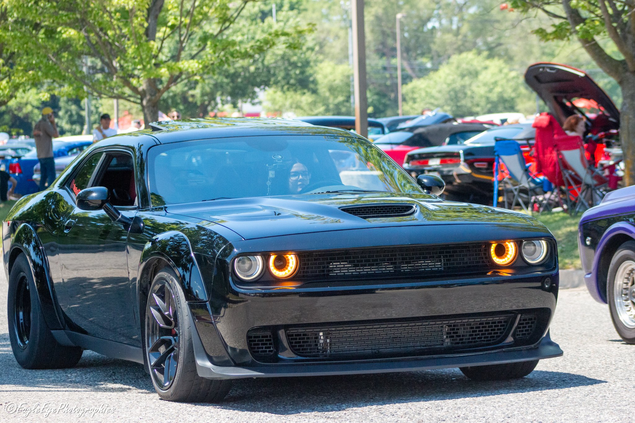 A shiny black sports car with orange headlights is parked outdoors at a car show. There are trees and multiple other vehicles in the background, along with people viewing the cars and a person with a dog. The car has a sleek design and reflective sur