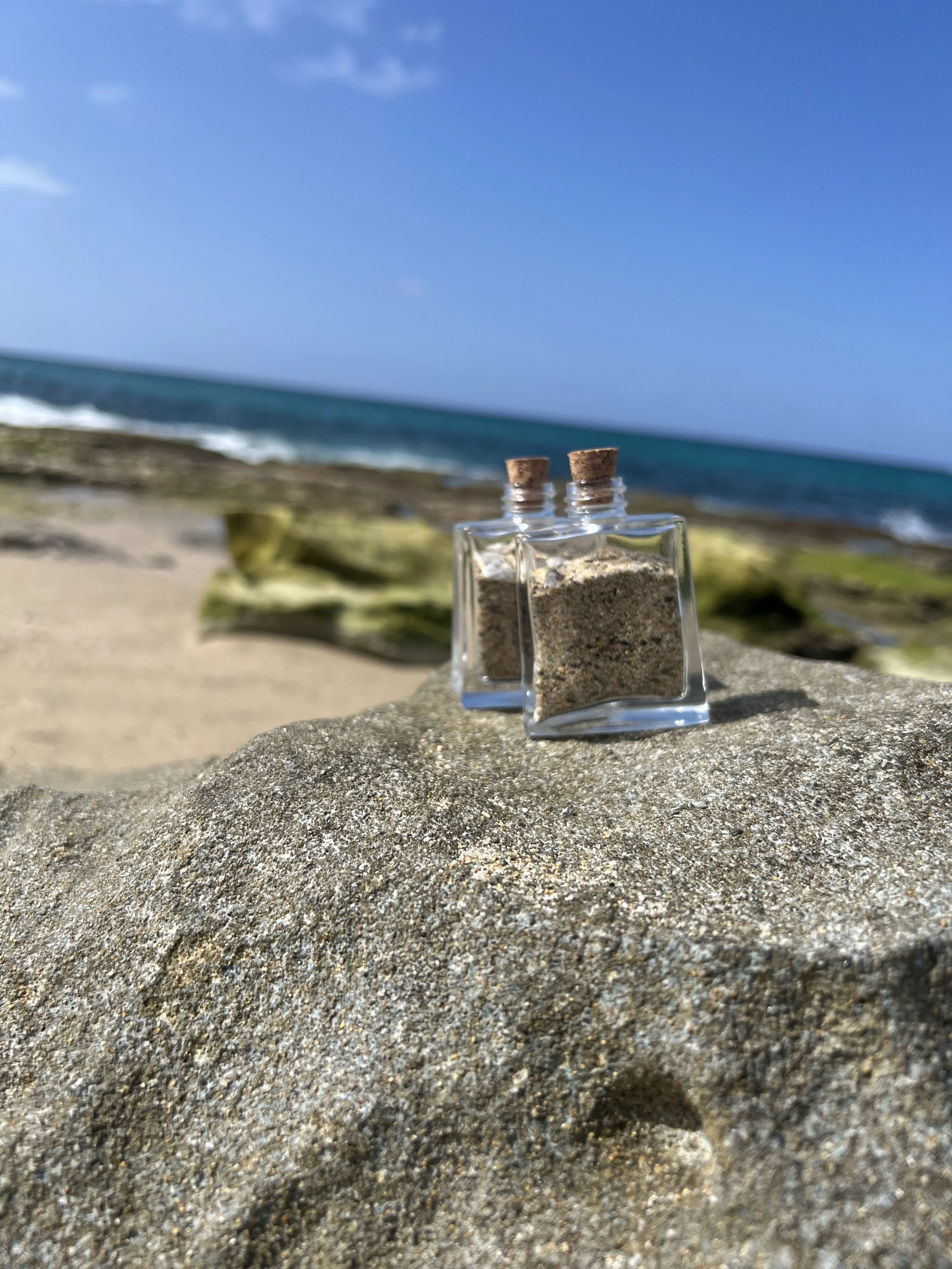 Close-up of two small glass bottles with cork stoppers containing sand, on a sandy beach with rocks, ocean waves, and a partly cloudy blue sky in the background.