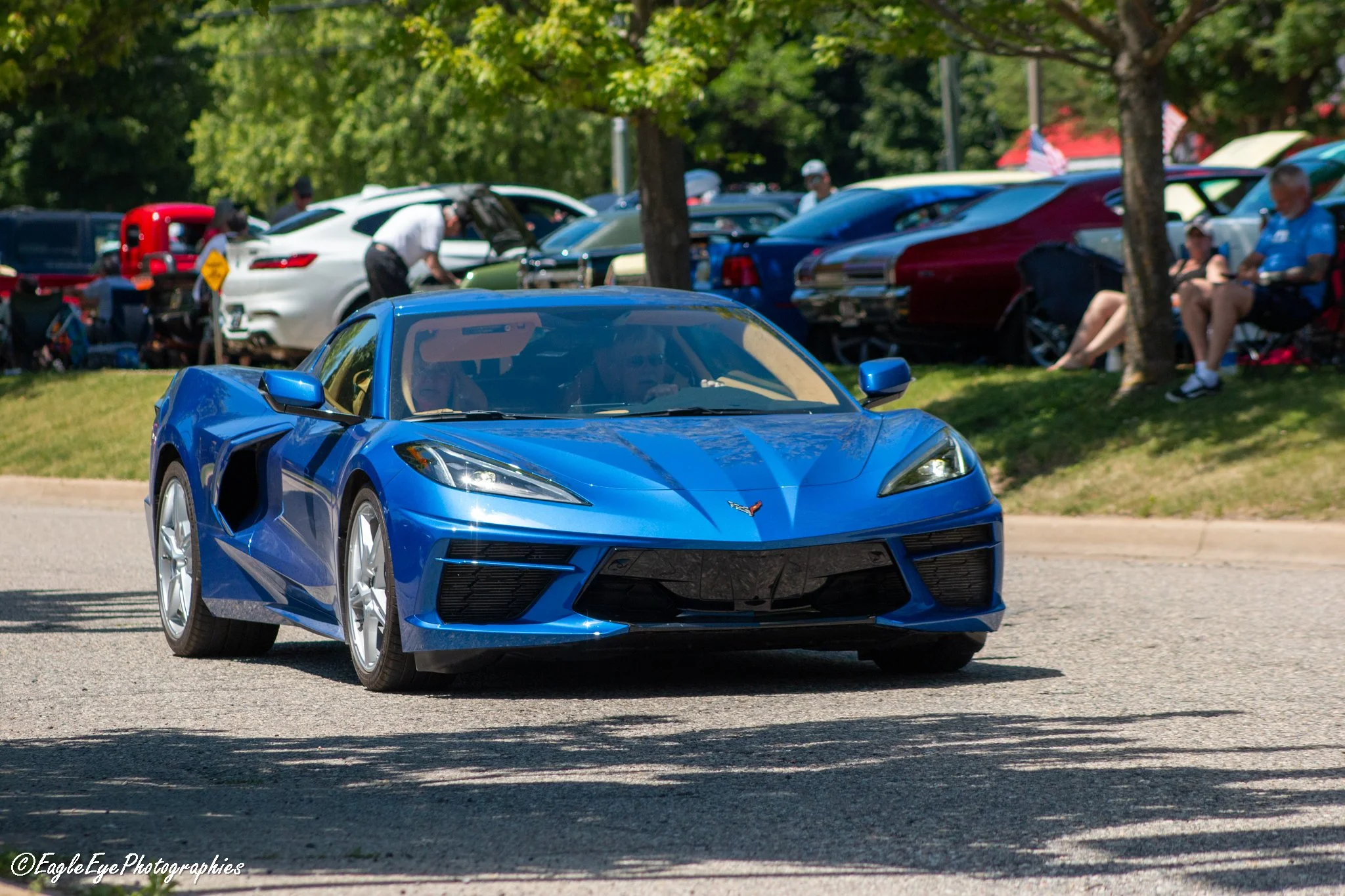 A vibrant blue sports car parked at a car show with other vehicles and people in the background under green trees.