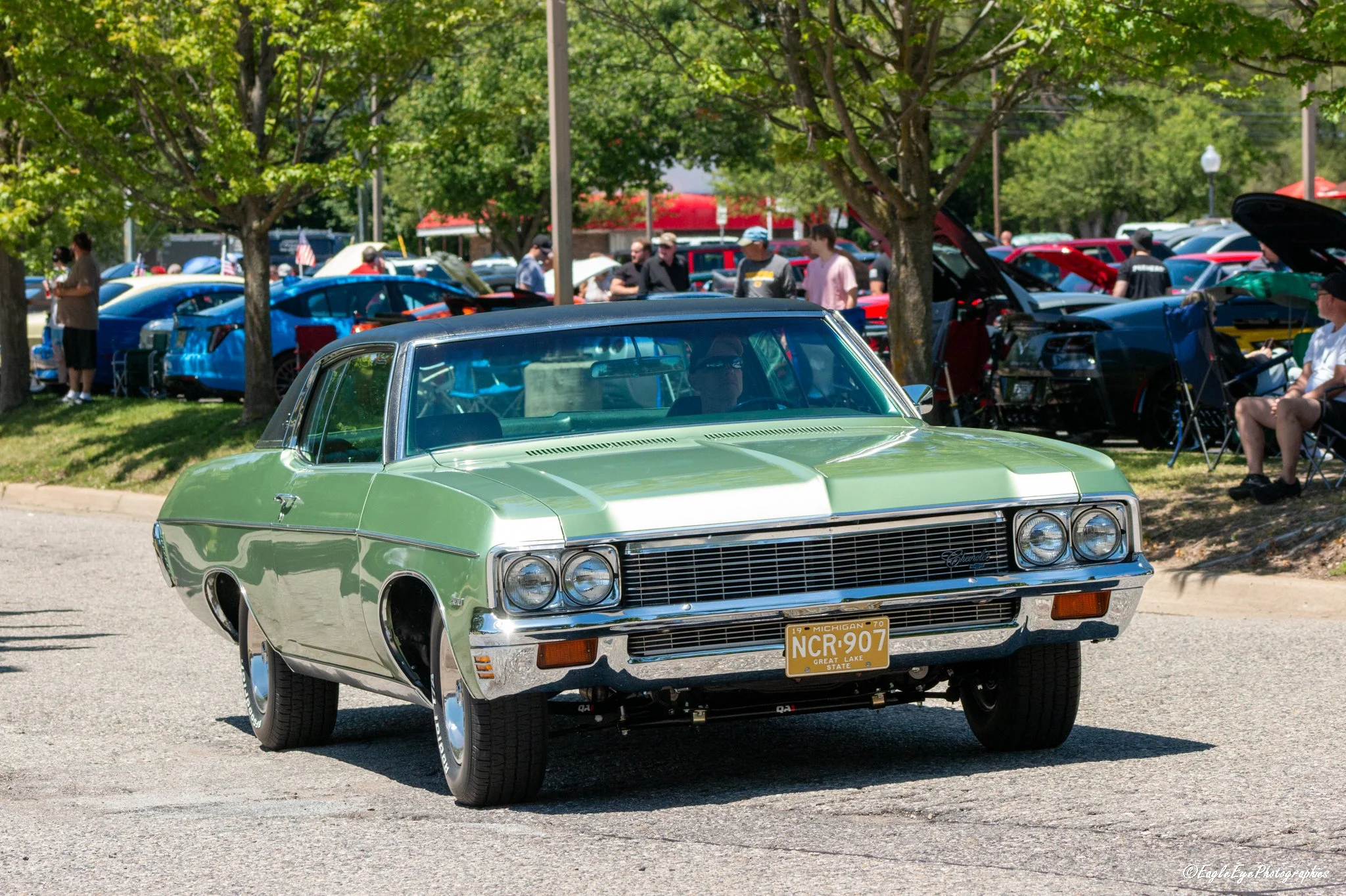A vintage green Cadillac with Michigan license plate NCR907 parked in front of a crowd at a car show under trees.