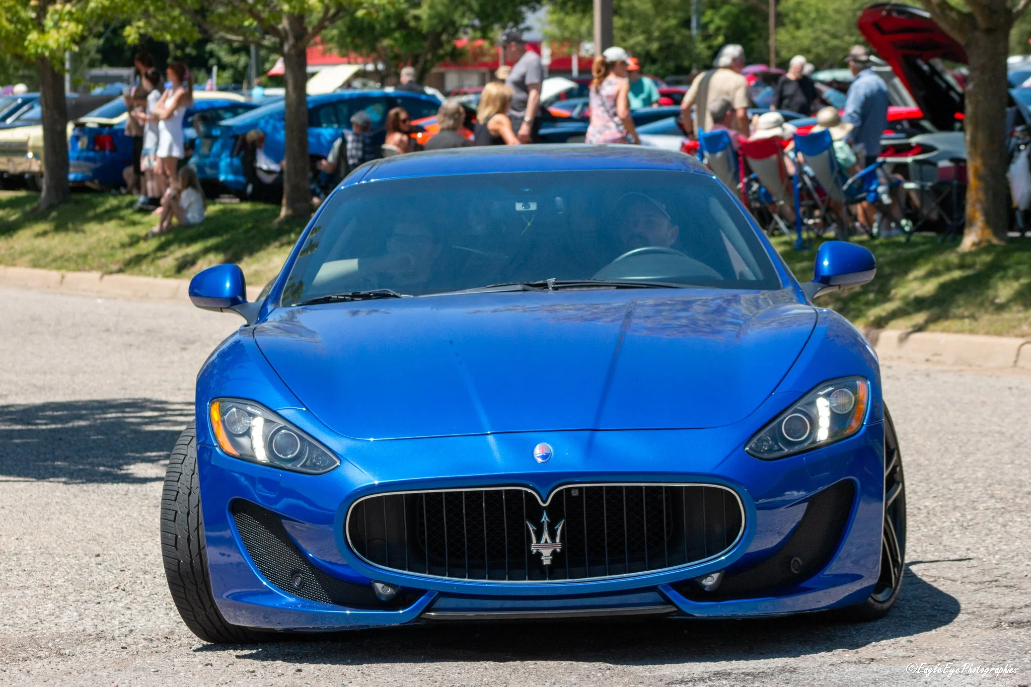 A bright blue sports car with a Maserati logo on the front grille parked on the street. In the background, there is a crowd of people, some sitting, standing, and walking near cars, with many parked cars and trees visible in a park-like setting.